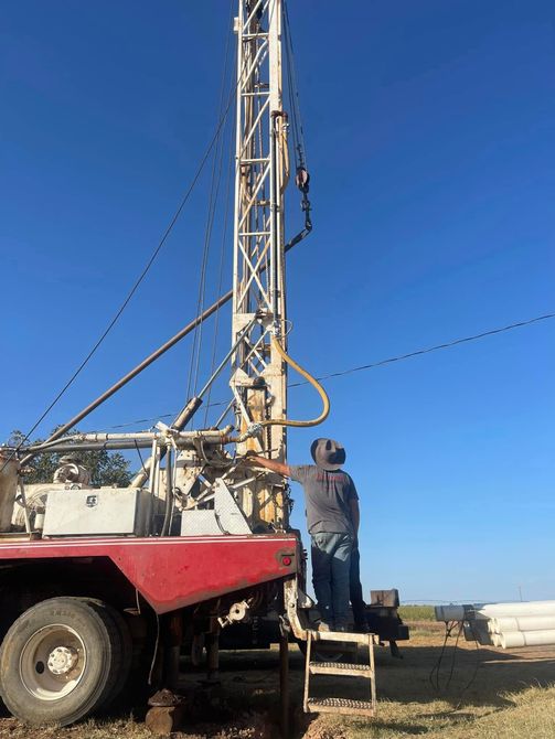 Worker on a red drilling rig beside a tall metal mast under a clear blue sky.