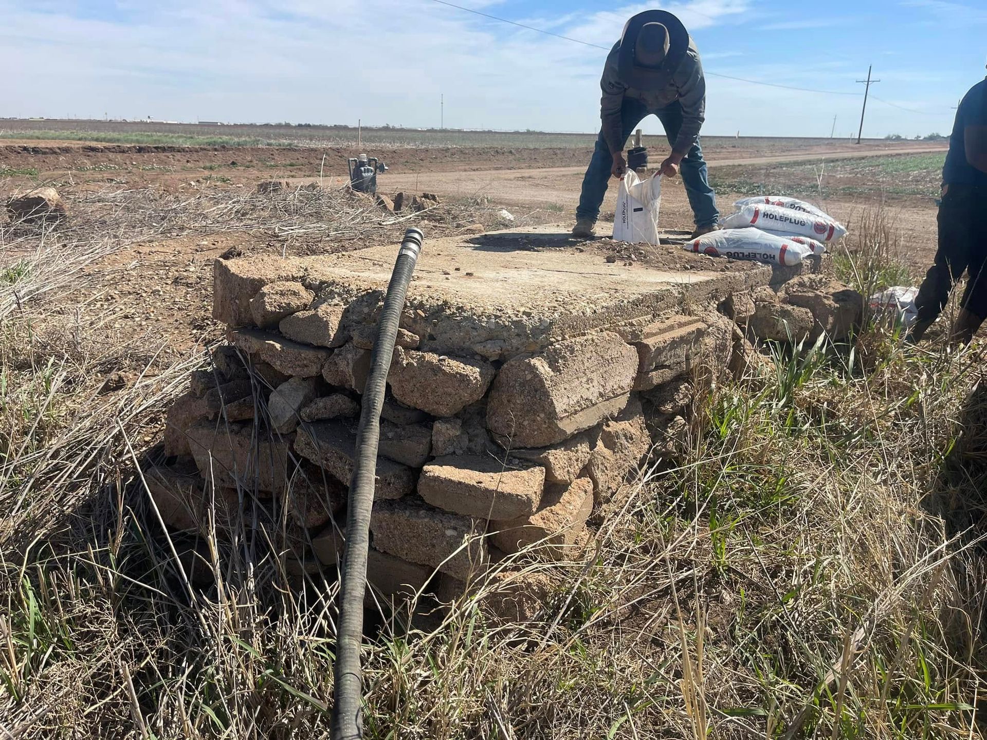 Workers kneel by a stacked stone cairn in a dry field, with a hose draped over it.