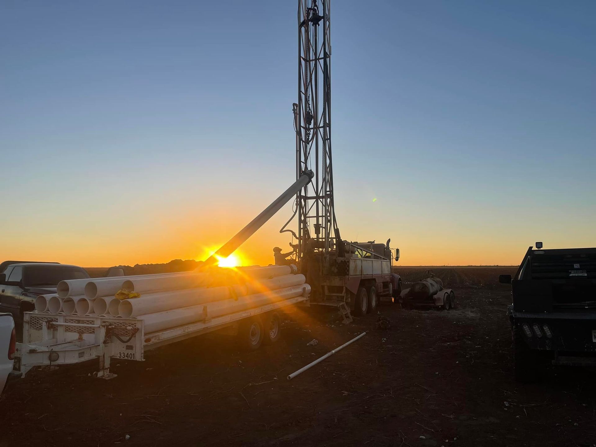 Oil rig silhouetted at sunset with drilling equipment and vehicles on a flat landscape