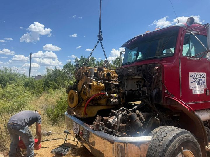 Worker using a crane to lift an engine into a red semi-truck in a field