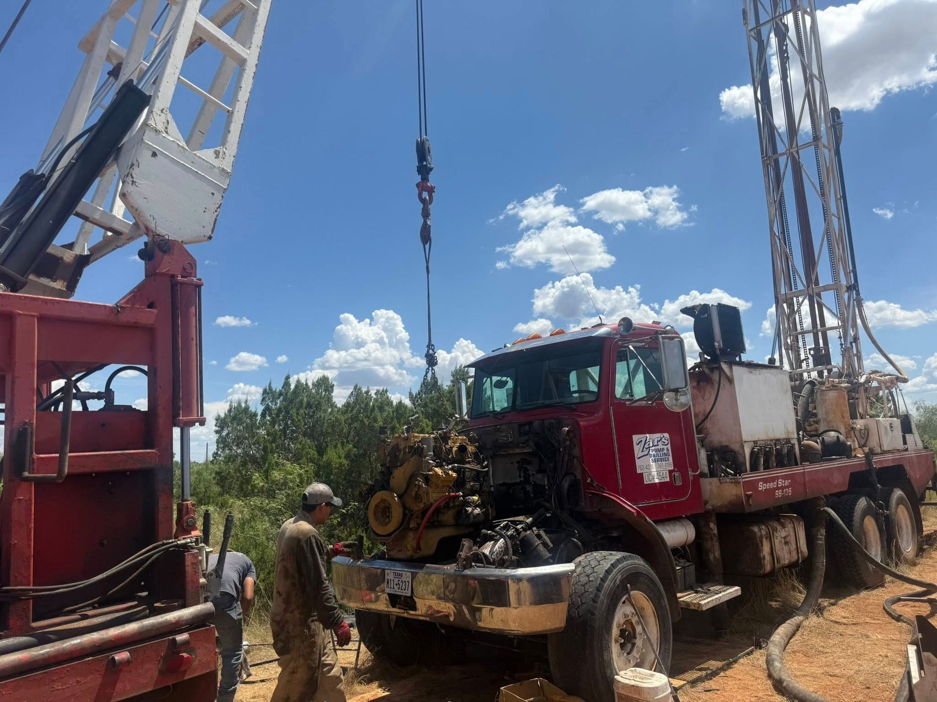 Red drilling truck at a well site with workers and tall rig equipment under a blue sky