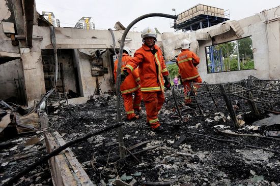 Firefighters in orange gear inspect a charred building after a fire.