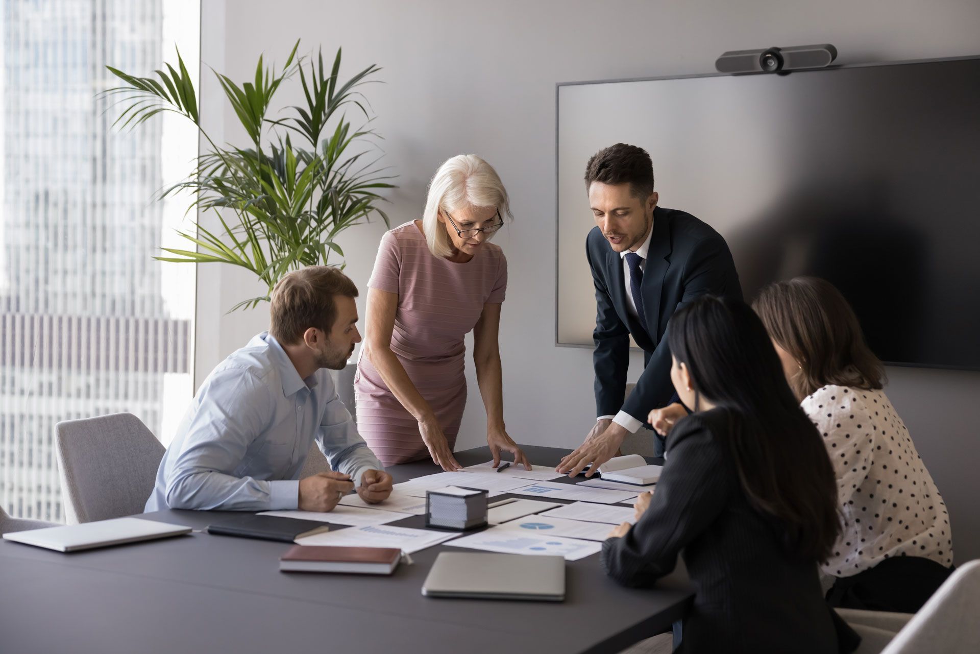 Team meeting around documents in a modern office, with a laptop, papers, and a wall screen.