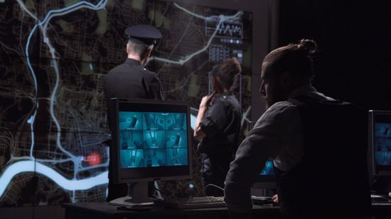 People monitoring blue computer screens in a dim surveillance room, with one standing near a glass wall.