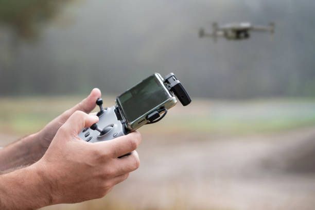 Hands holding a drone controller with a smartphone, with a flying drone blurred in the background.