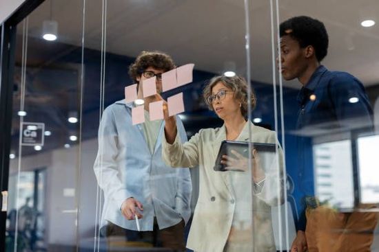 Three coworkers discussing sticky notes on a glass wall in a modern office