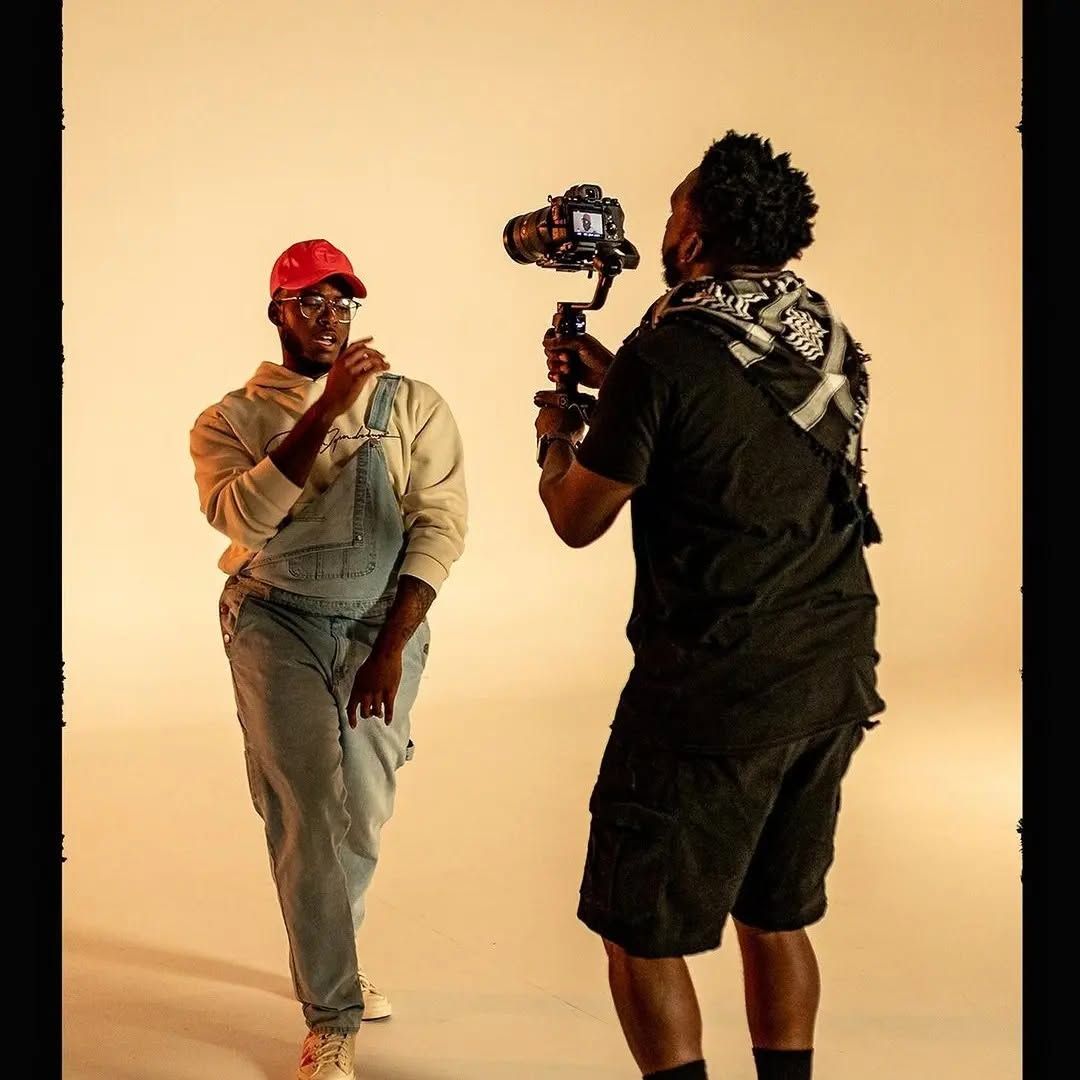 Two people filming a dance pose in a studio, one holding a camera rig toward the other.