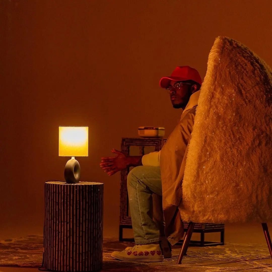 Person in red cap sits by a glowing lamp beside a textured chair in a warm orange room