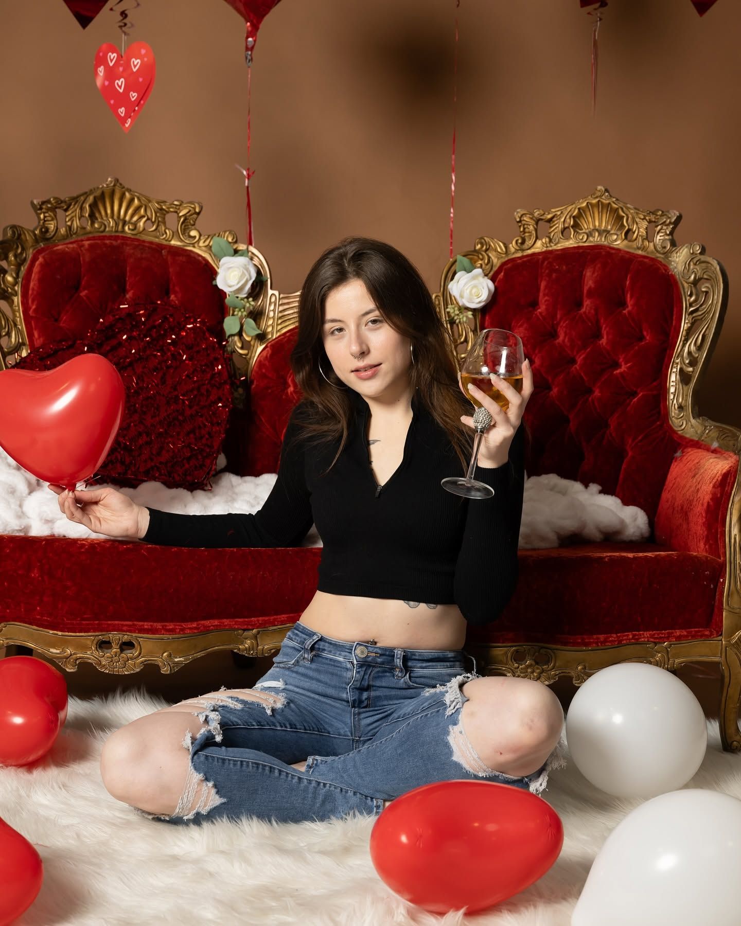 Woman in black top and ripped jeans sitting amid red and white balloons on ornate red chairs, holding a drink