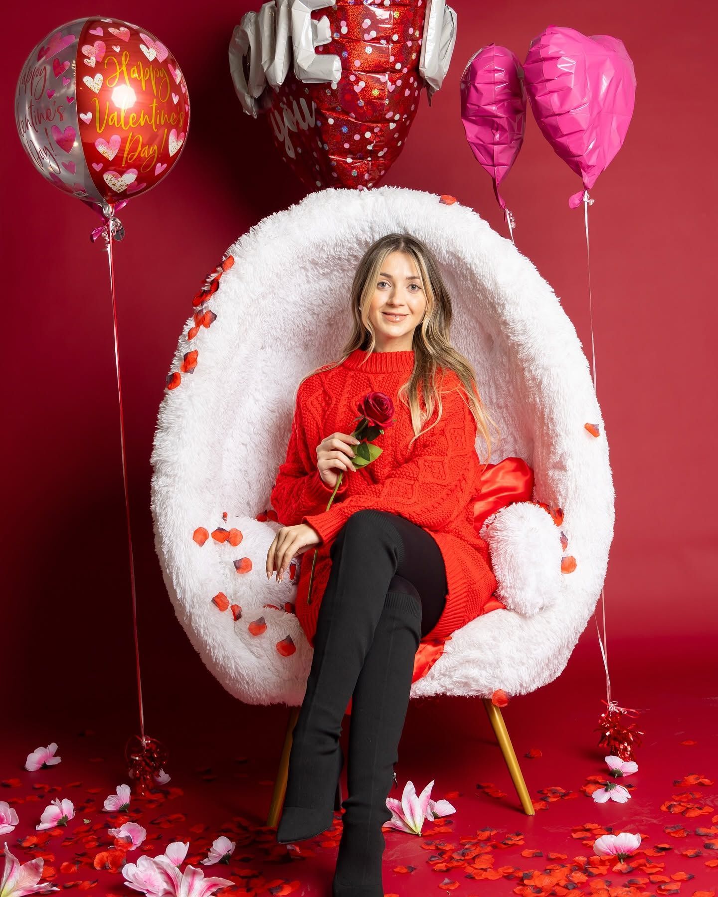 Woman in red sweater sitting in a white chair with balloons and rose petals on a red backdrop.
