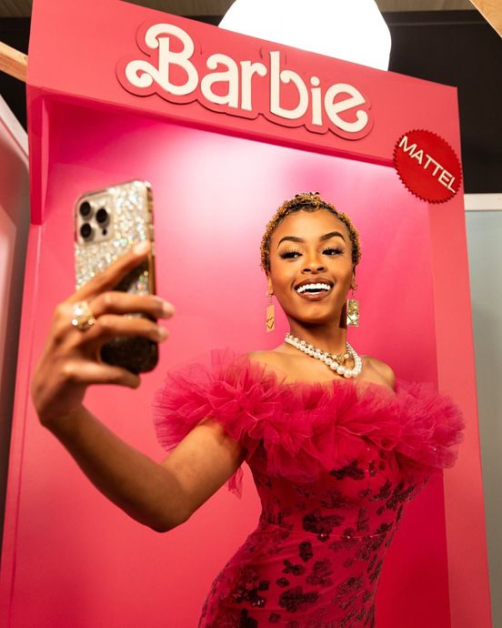 Woman in a bright pink Barbie display taking a selfie, wearing a pink dress and jewelry.