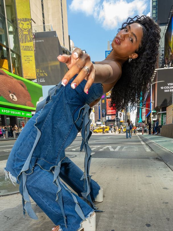 Woman in denim pose on a city street, reaching toward the camera with a playful smile.