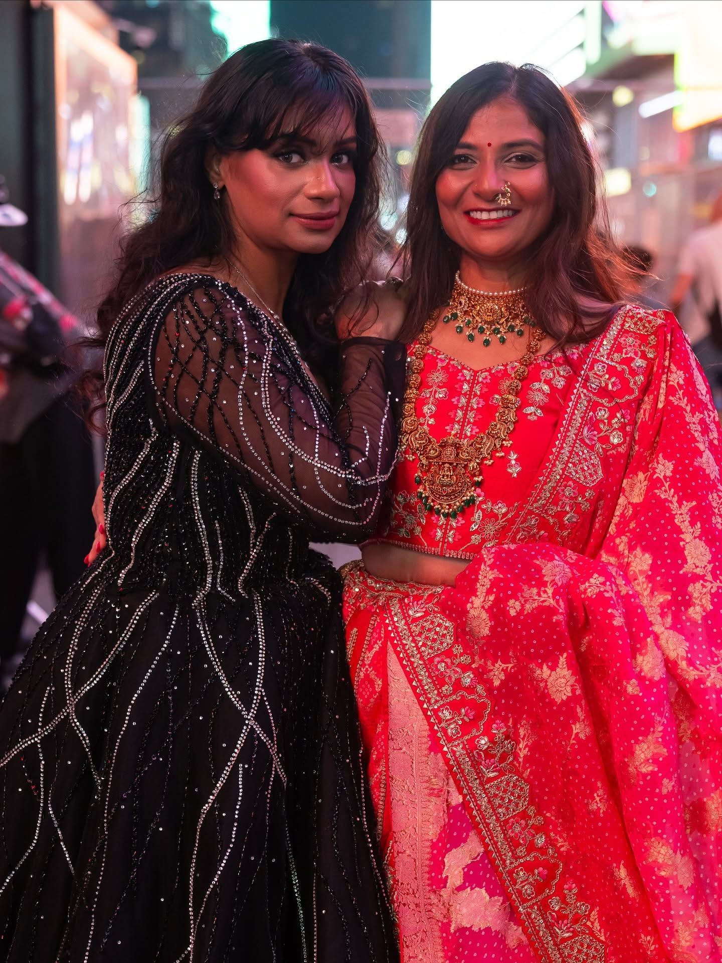 Two women posing together in festive black and red traditional outfits on a city street at night