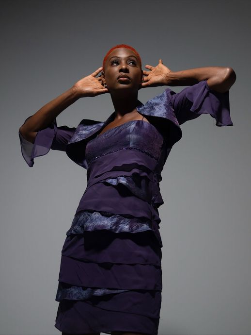 Model in a purple layered dress posing with hands near ears against a gray backdrop