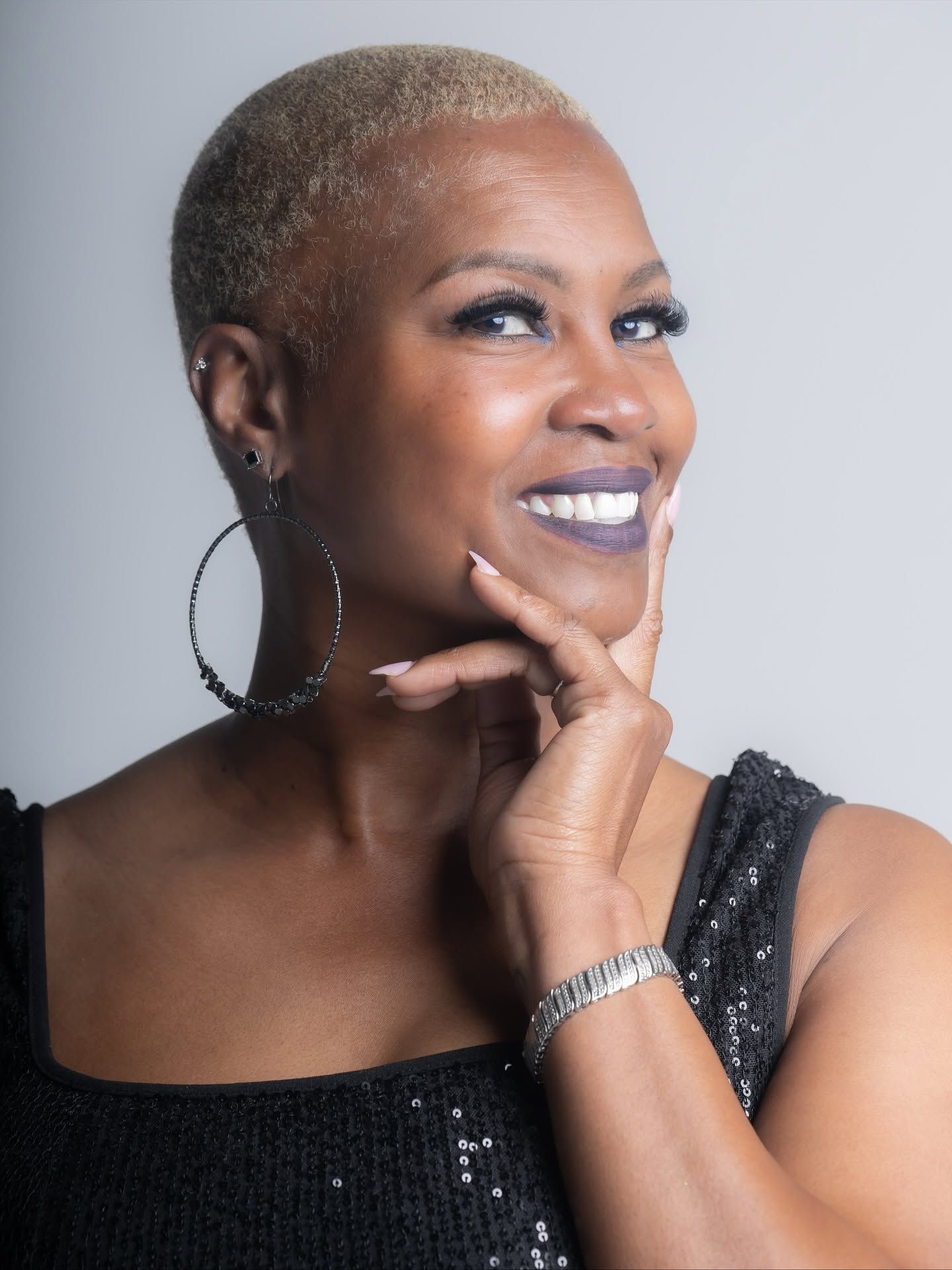 Smiling woman with short blonde hair, wearing a black dress and silver jewelry, posing against a gray background