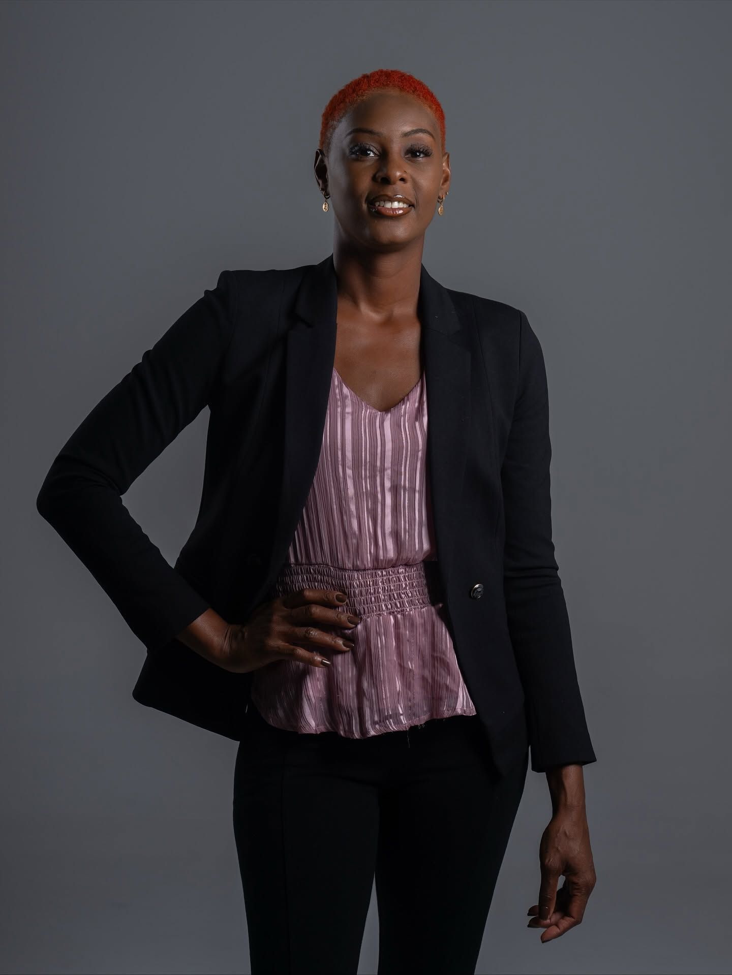 Person in a black blazer and pink blouse poses against a gray studio backdrop.