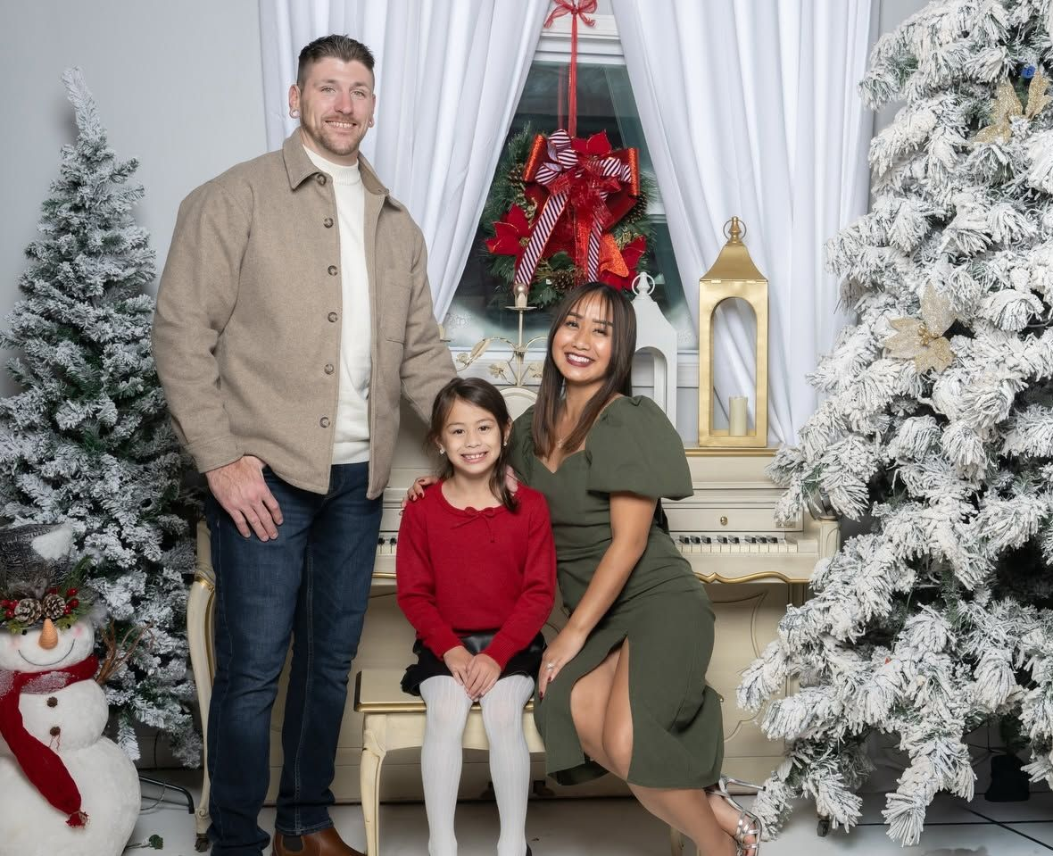 Family posing by a decorated Christmas fireplace with snow-covered trees and a red wreath.