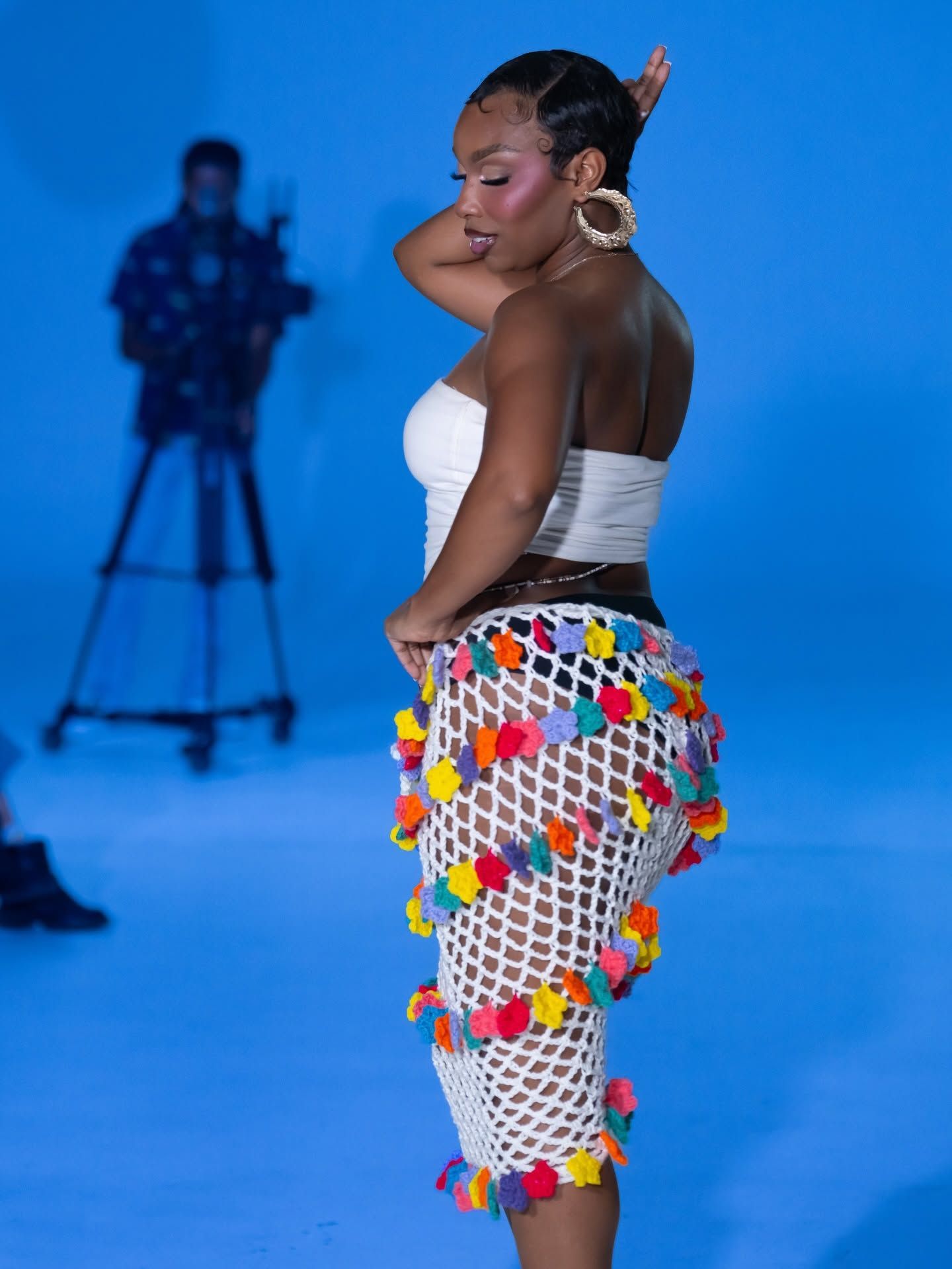 Woman in white top and colorful skirt poses on a blue studio set with a camera in the background