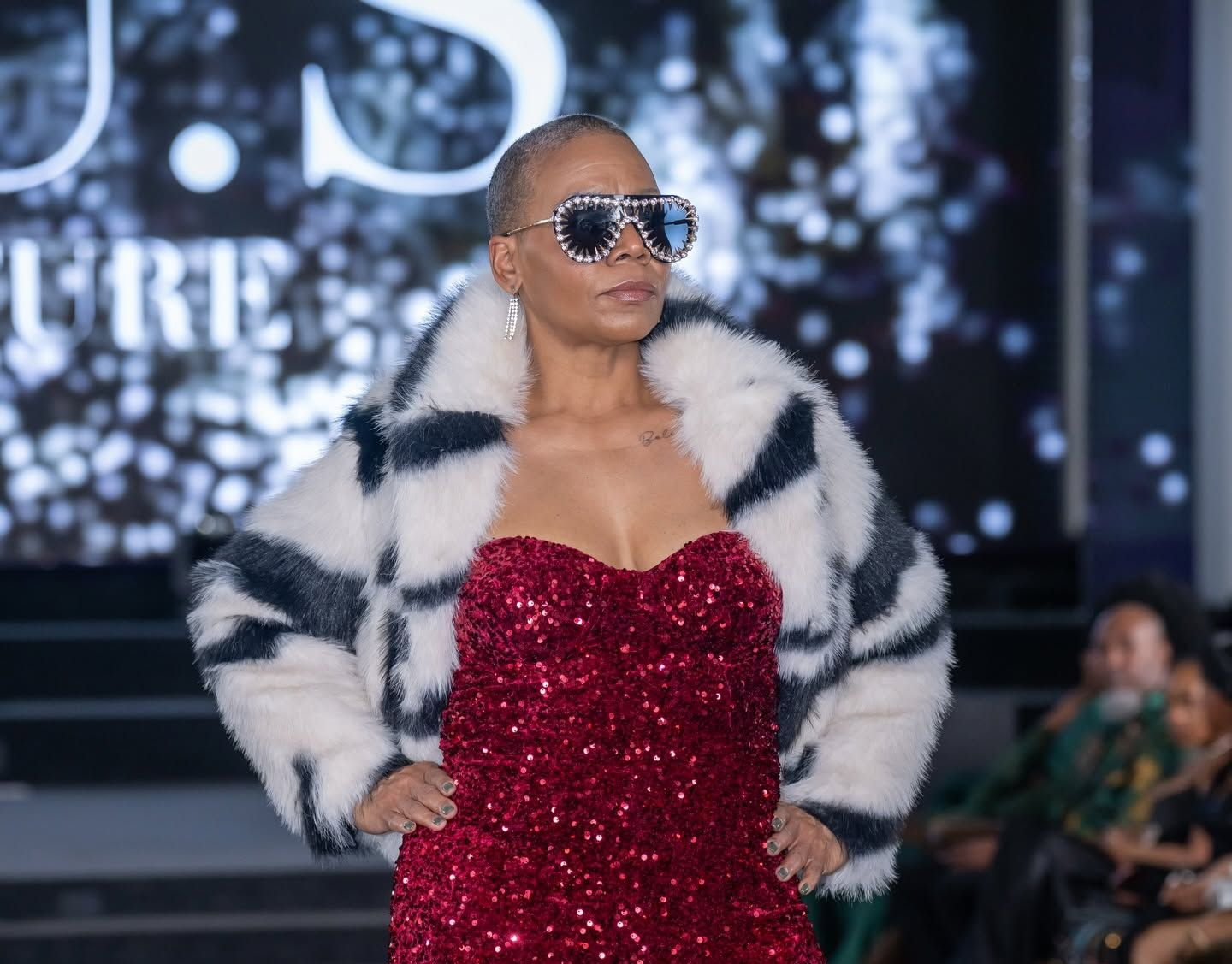 Woman in a red sequined dress and striped fur coat, wearing sunglasses, on a runway with a blurred backdrop
