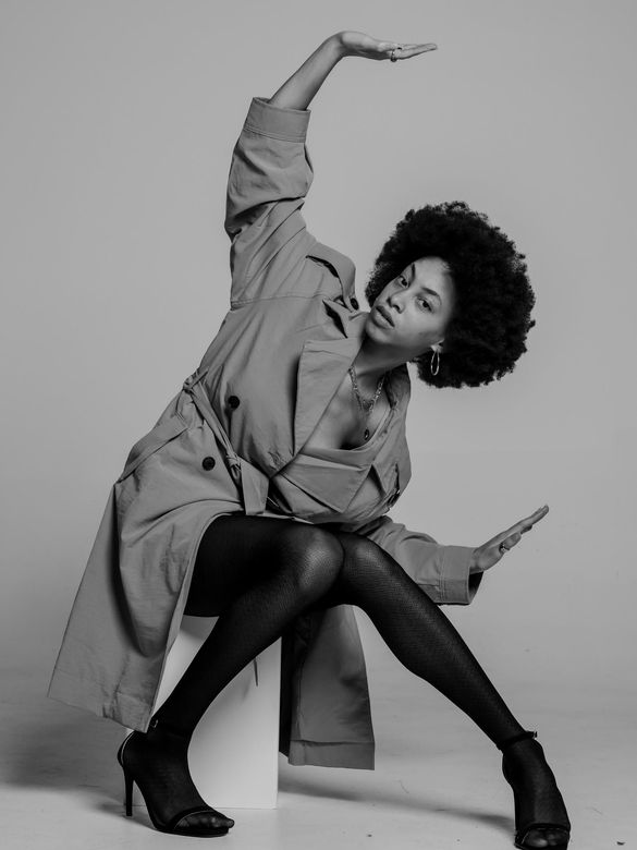 Person in a coat and heels striking a dynamic seated pose on a plain studio backdrop.
