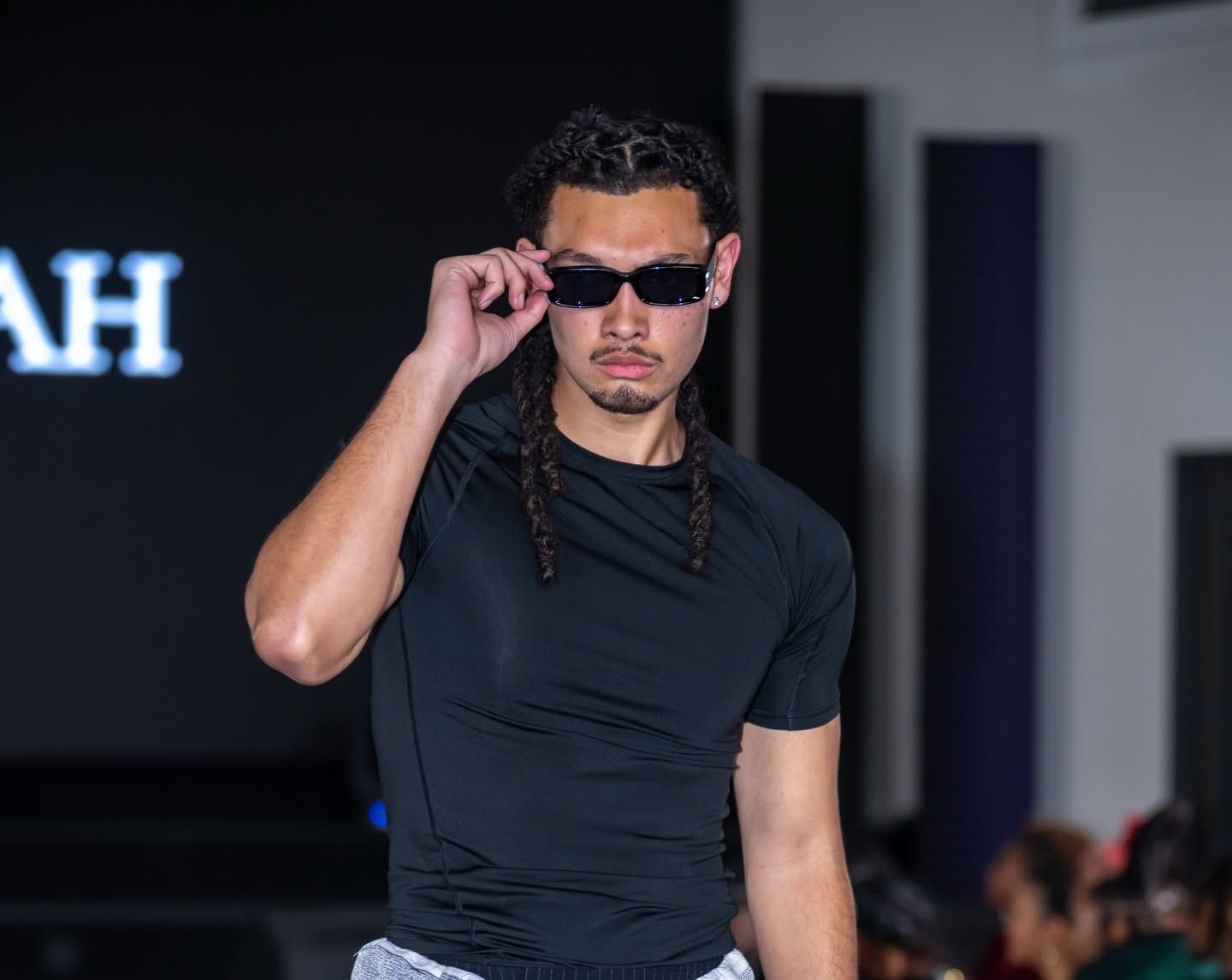 Man in black shirt adjusting sunglasses on a runway, with a dark stage background