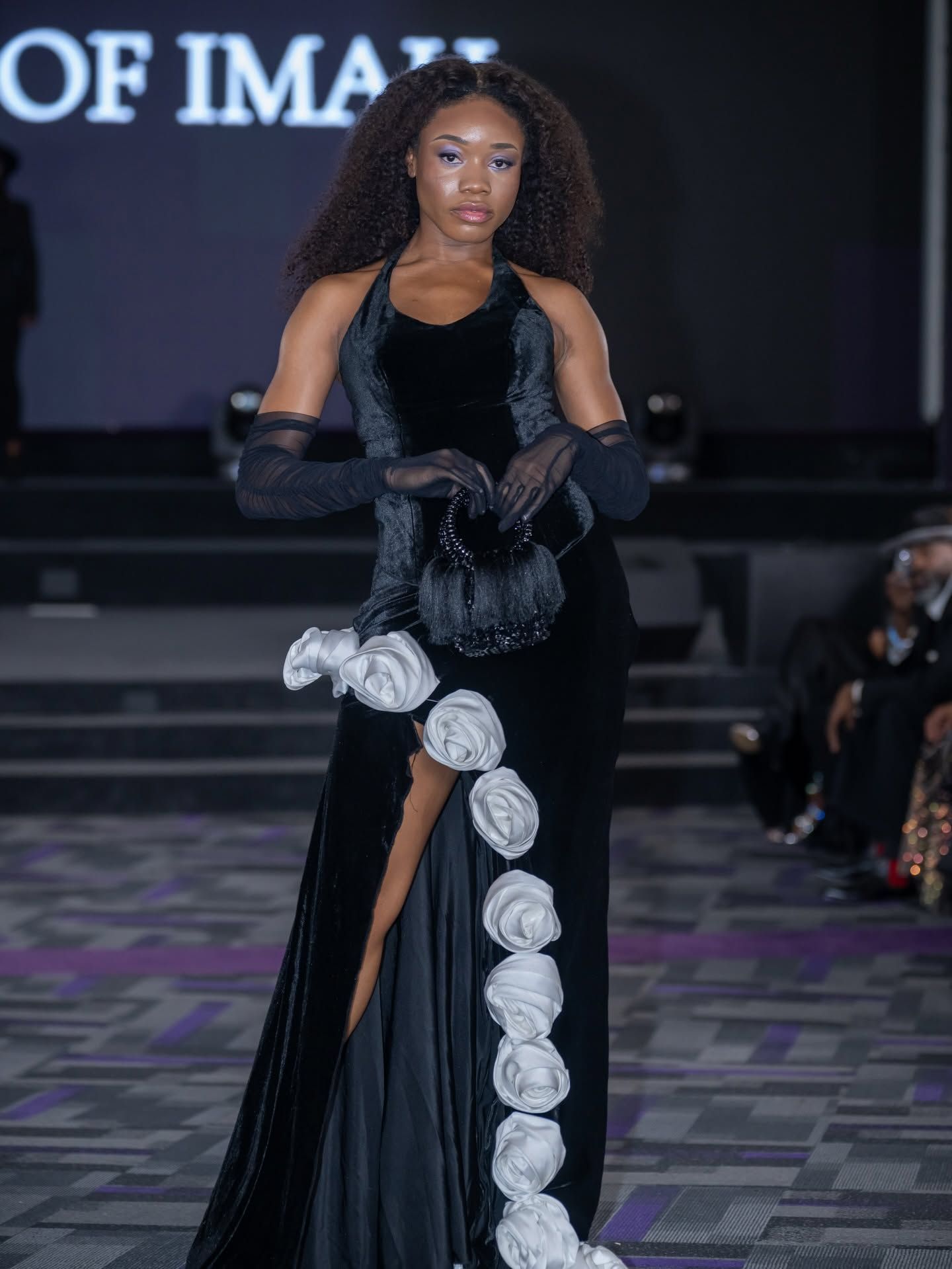 Model in a black evening gown with white floral trim walking a runway.