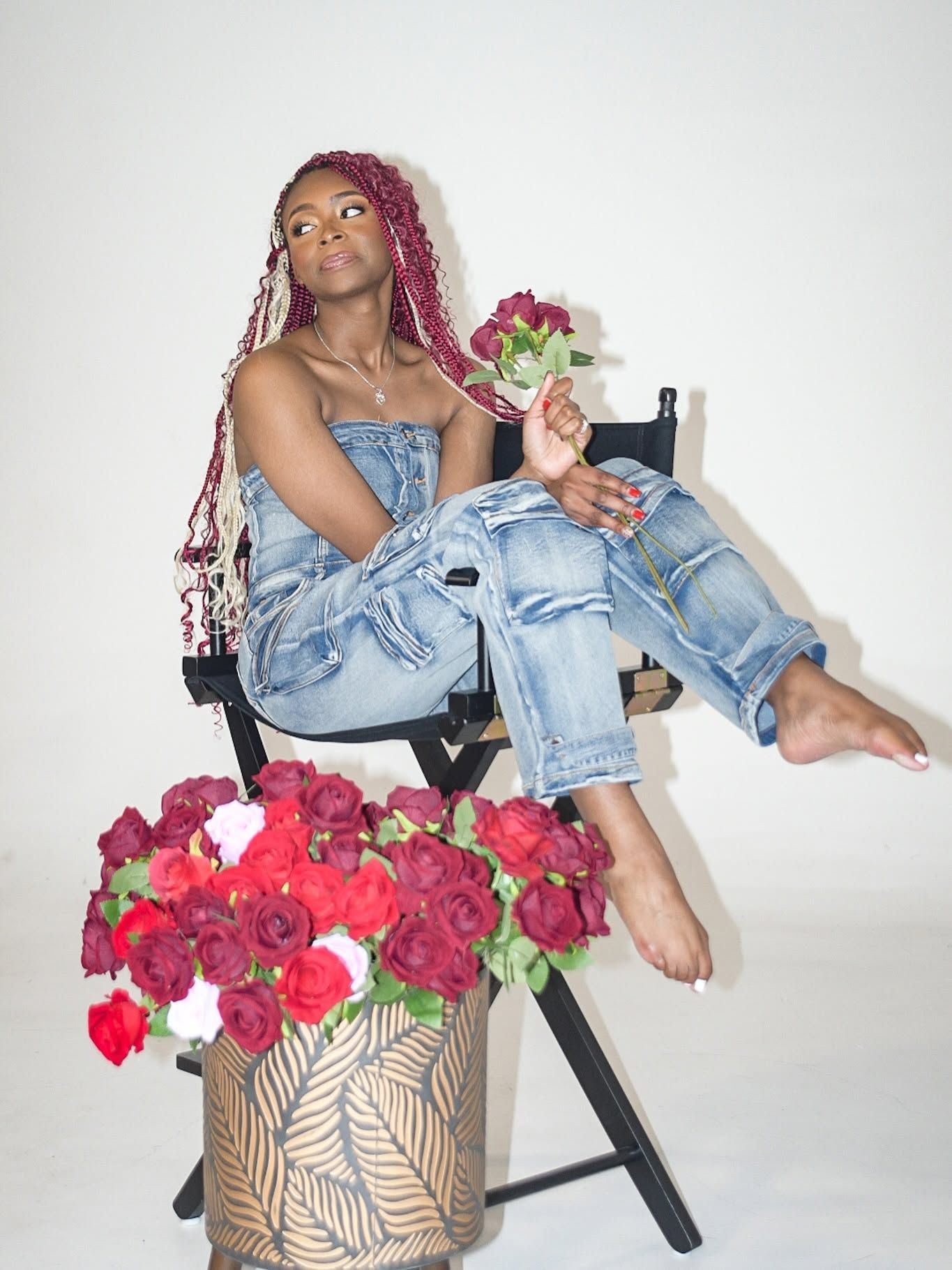 Person seated on a chair holding flowers, wearing a blue denim outfit beside a vase of red and pink roses.