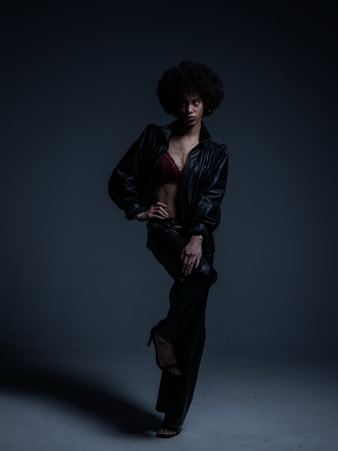 Person in a black jacket and pants poses against a dark studio backdrop.