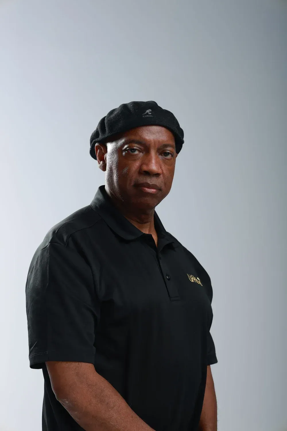 Man in black shirt and beret standing against a plain gray background, facing the camera.