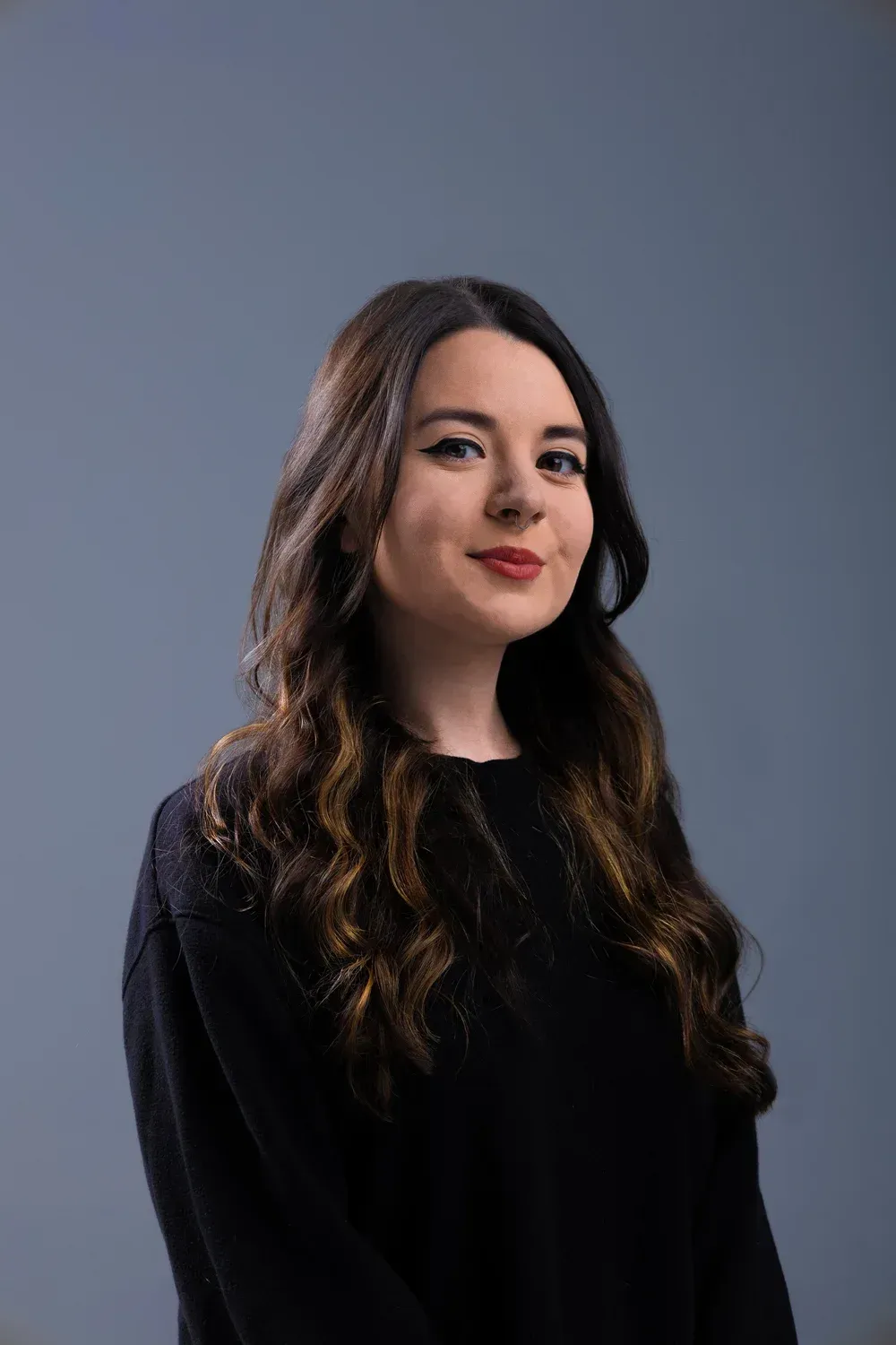 Woman with long dark hair in a black top against a gray studio background, smiling softly