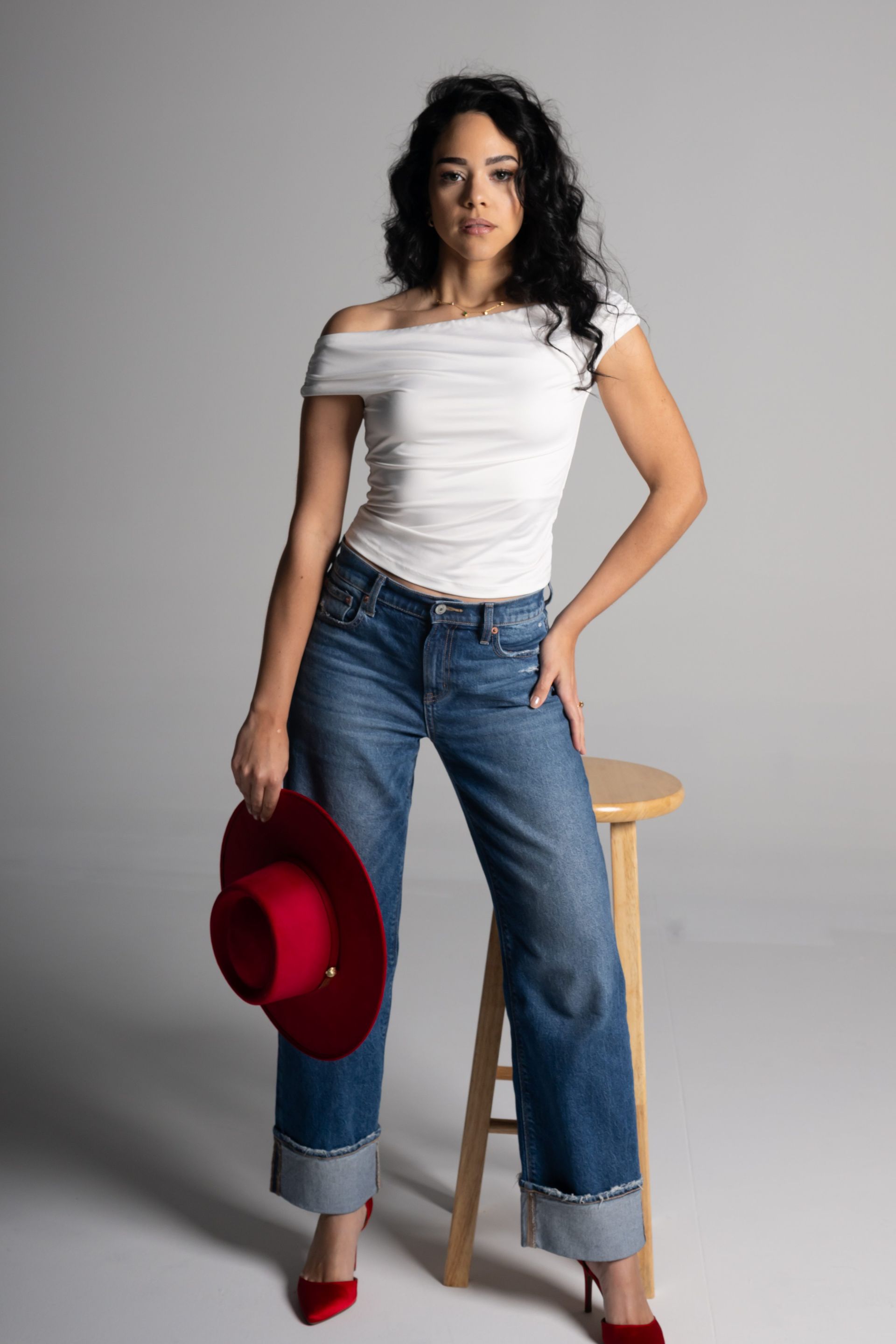 Woman in white top and jeans standing with a red hat beside a wooden stool against a gray backdrop