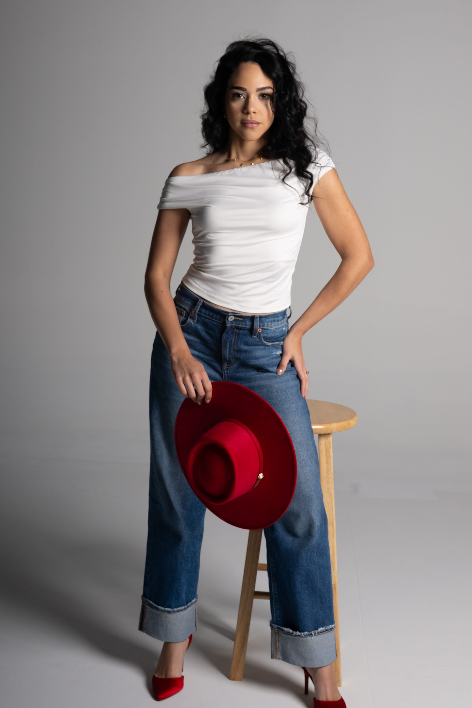 Woman in white top and jeans holding a red hat beside a wooden stool against a gray backdrop
