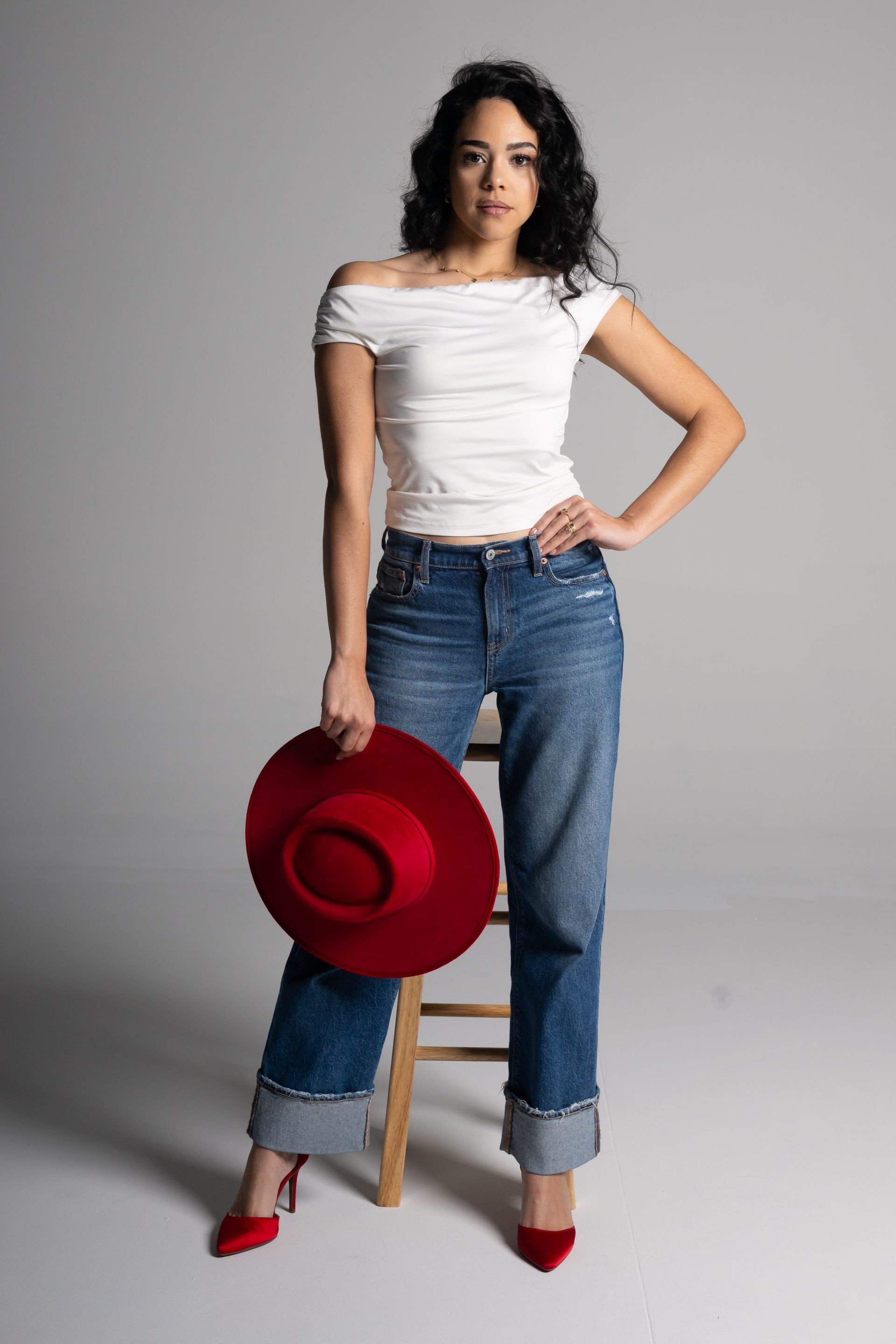 Woman in white top and jeans holding a red hat, standing beside a stool on a gray background.