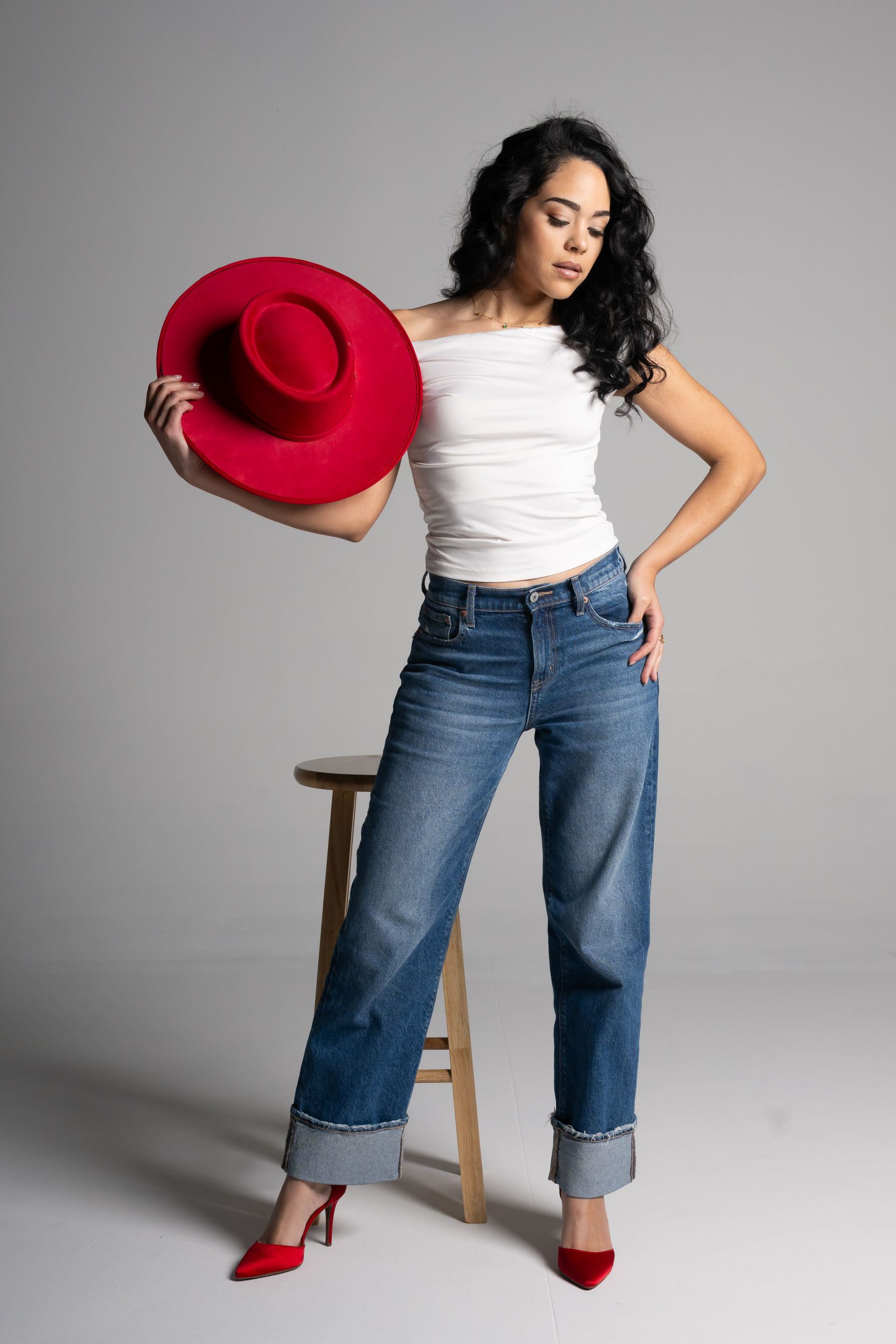 Woman in white top and jeans posing with a red hat against a gray studio backdrop