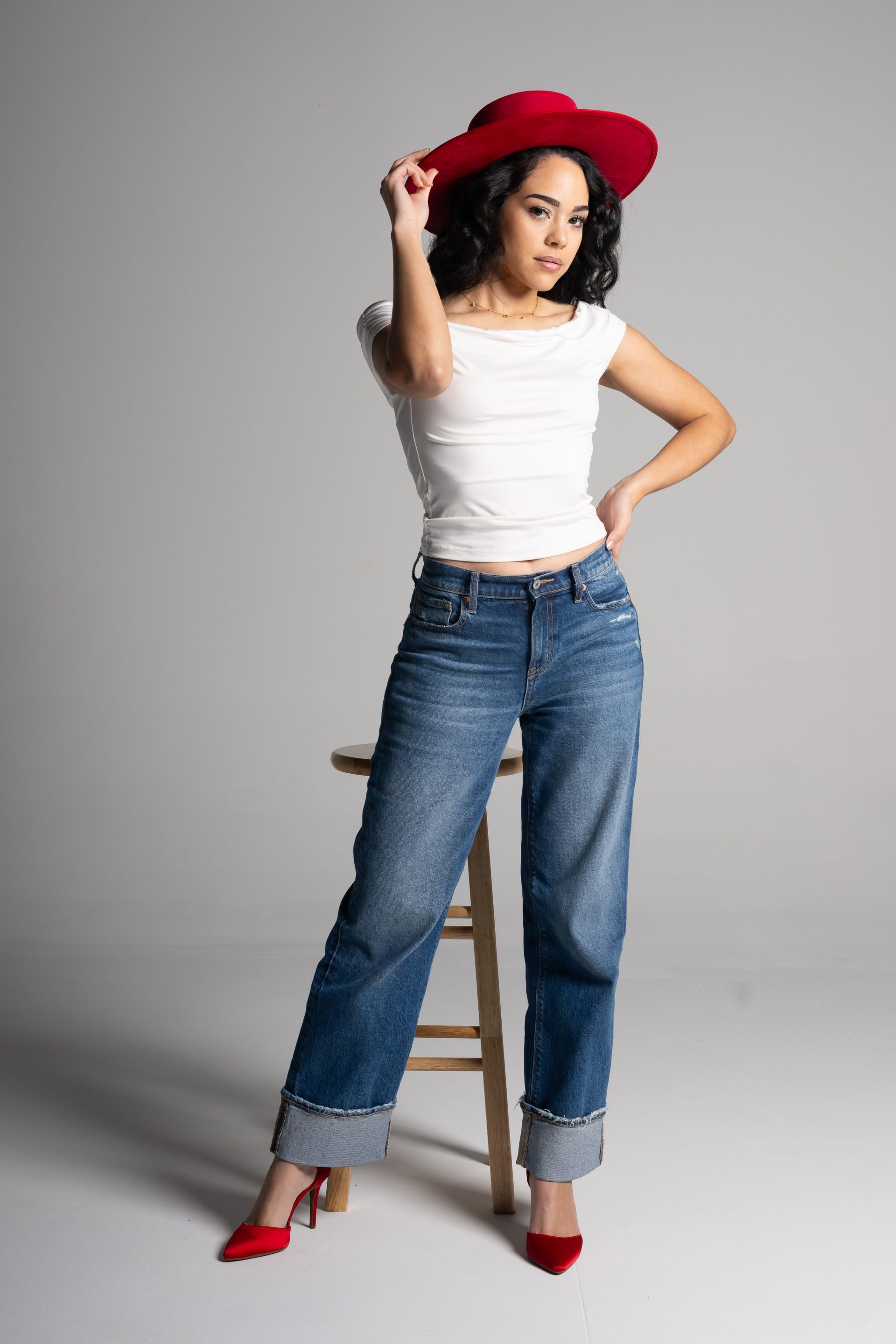 Woman in a red hat, white top, and jeans posing on a stool against a gray backdrop.