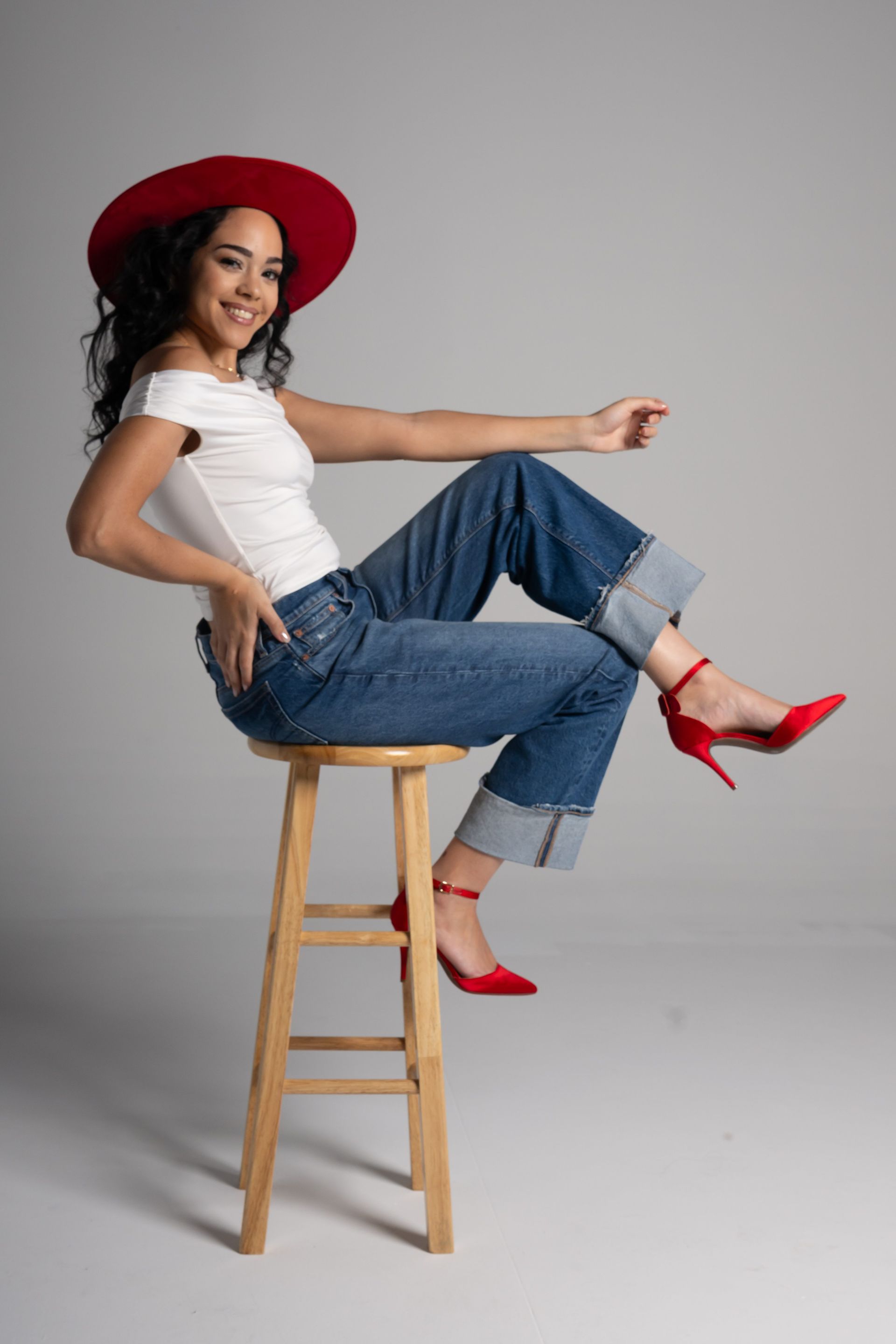 Woman in a red hat and red heels posing on a wooden stool against a gray backdrop.