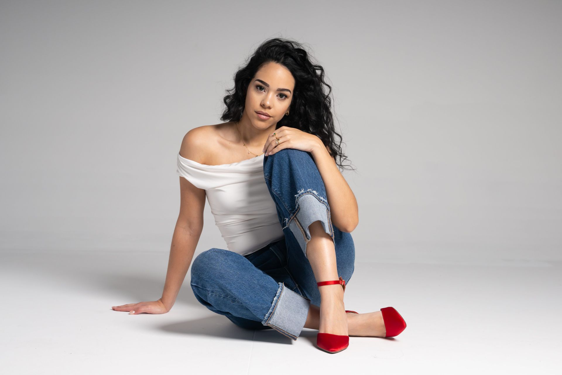 Woman seated on a studio floor wearing an off-shoulder white top, jeans, and red heels against a gray background