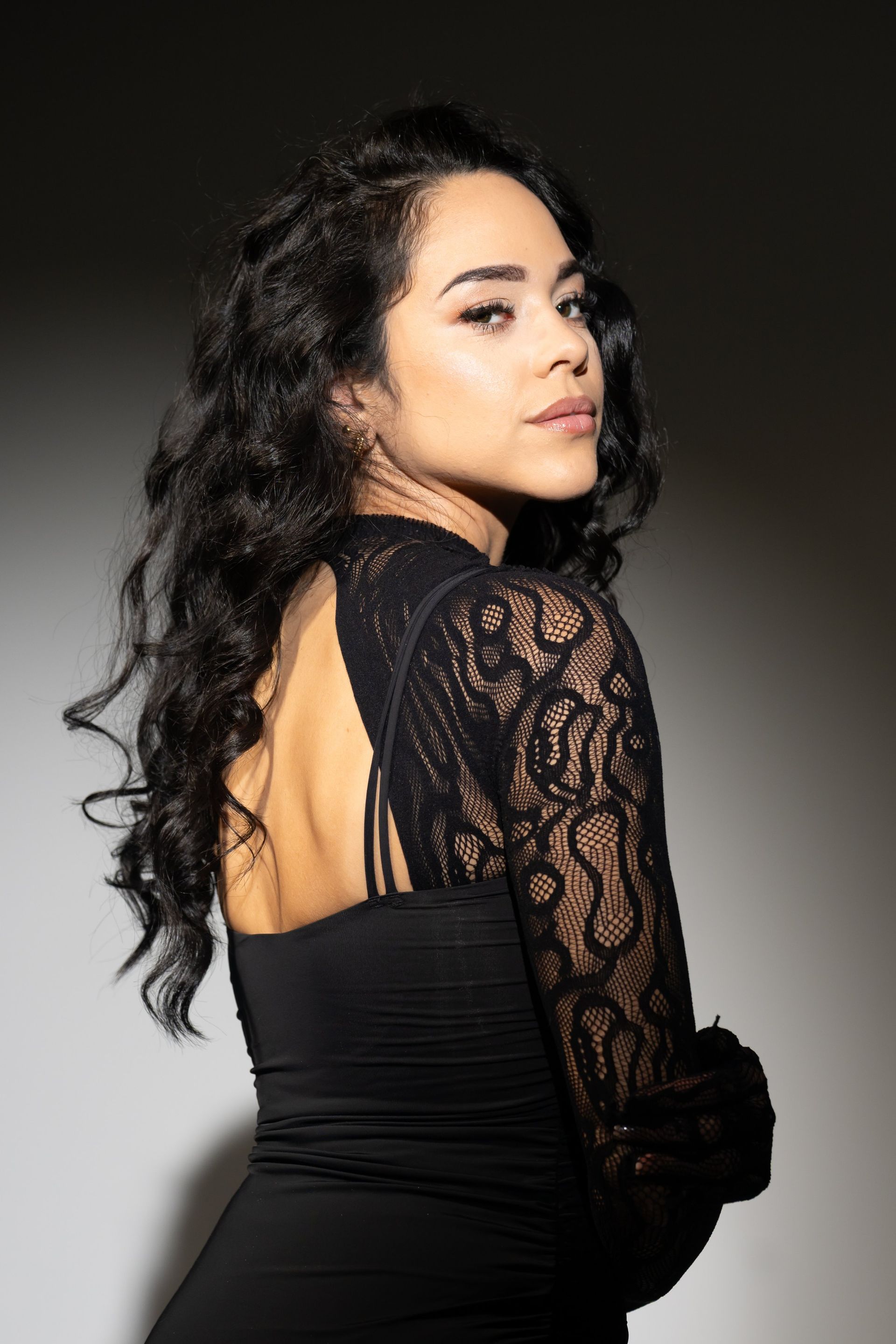 Woman in a black lace dress posing against a dark studio backdrop, looking over her shoulder.