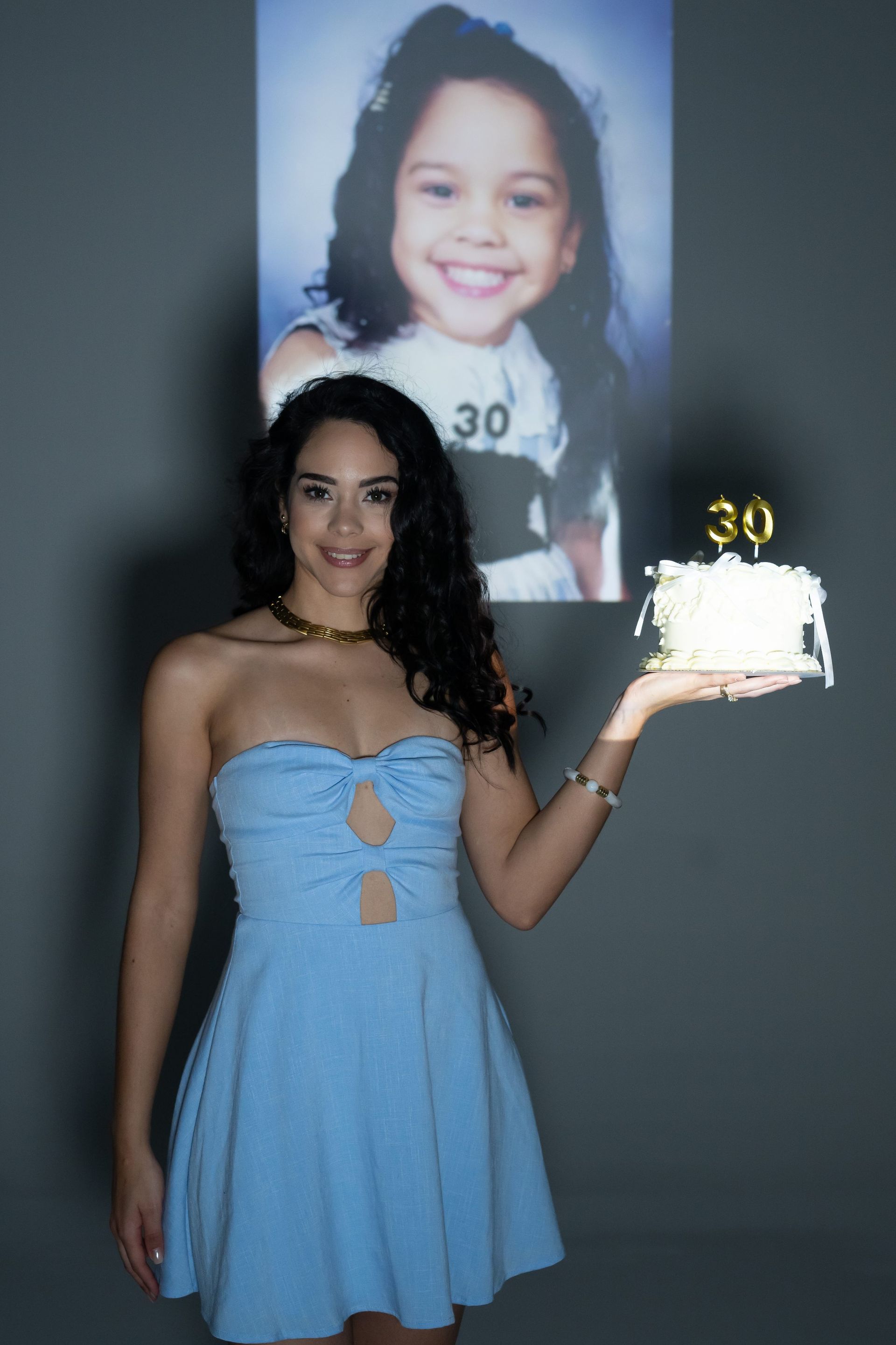 Woman in a light blue dress holding a small cake with a “30” candle, standing before a projected child photo.