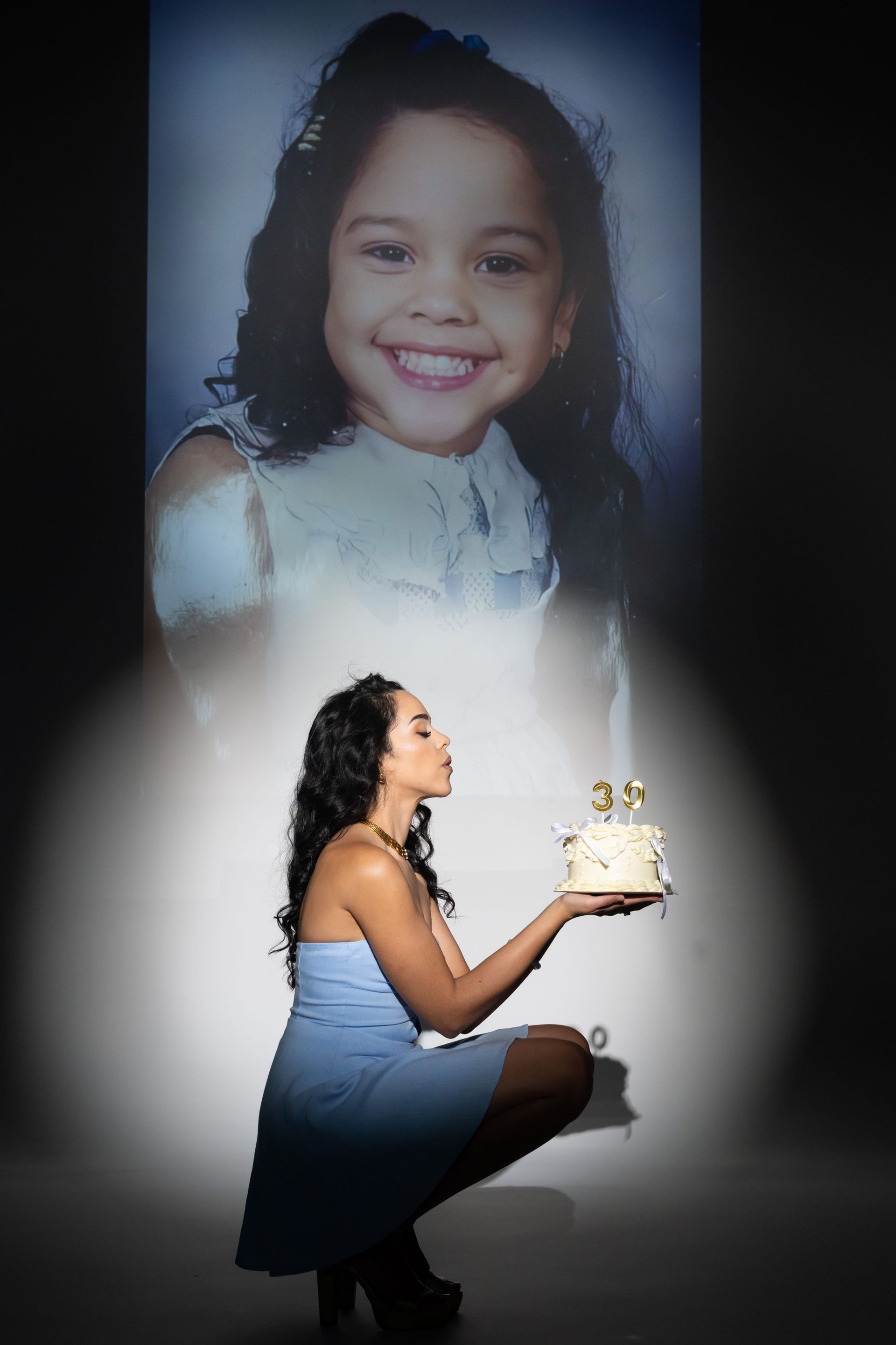 Woman kneeling with a birthday cake beneath a large portrait of a smiling child on a gray backdrop