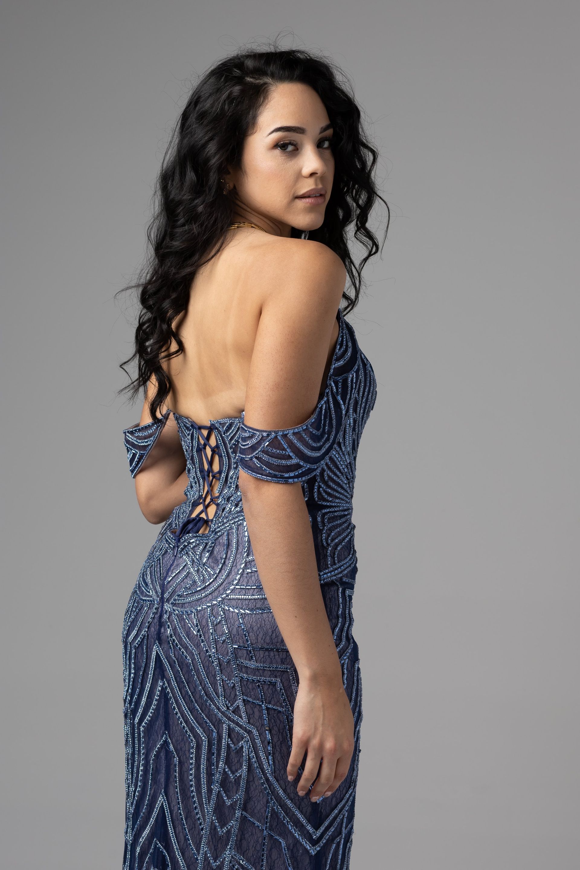 Woman in a blue patterned dress posing against a gray backdrop, looking over her shoulder.