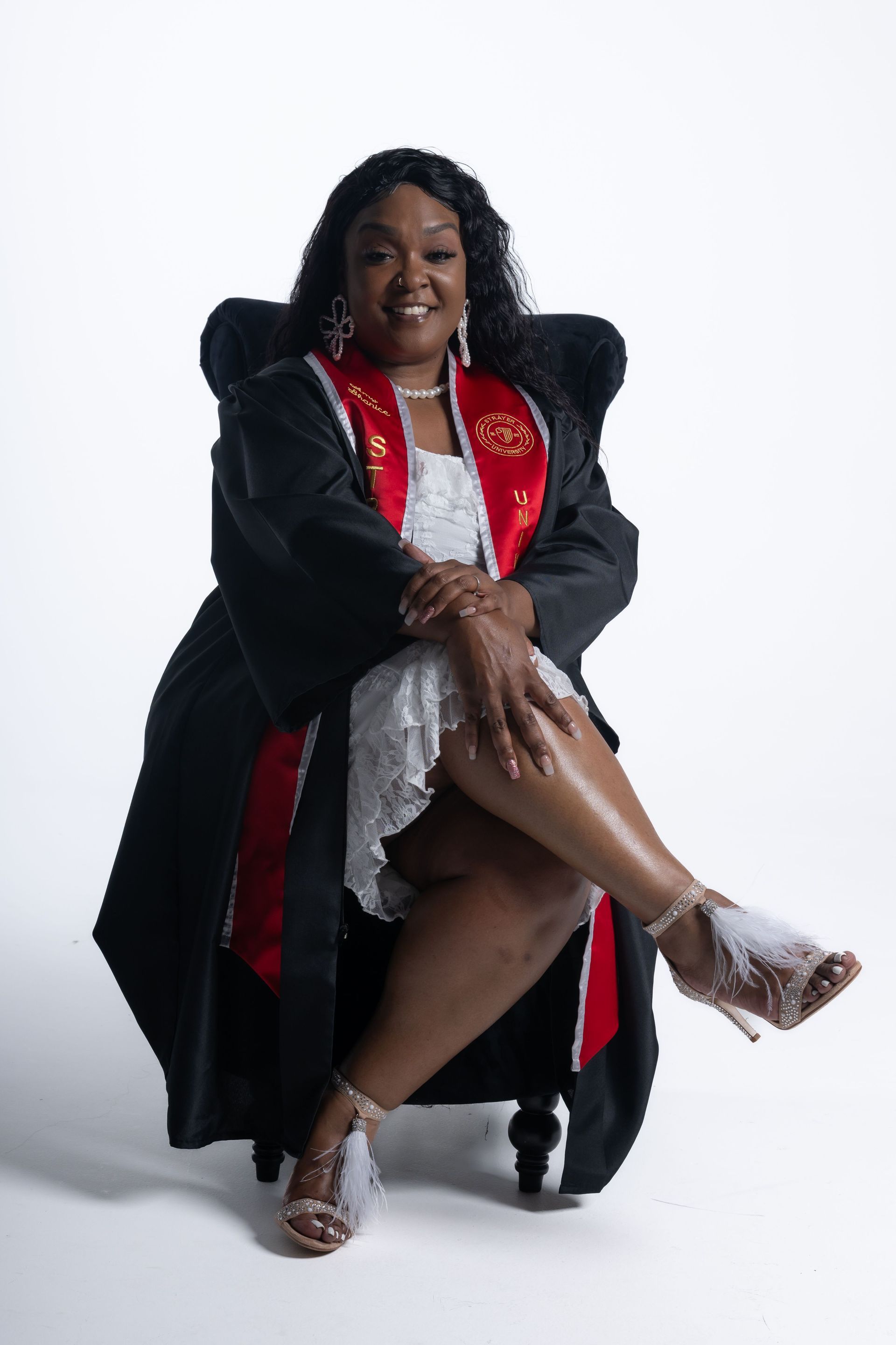 Person seated on a stool, wearing a black coat with red lining and silver outfit, smiling against a white backdrop