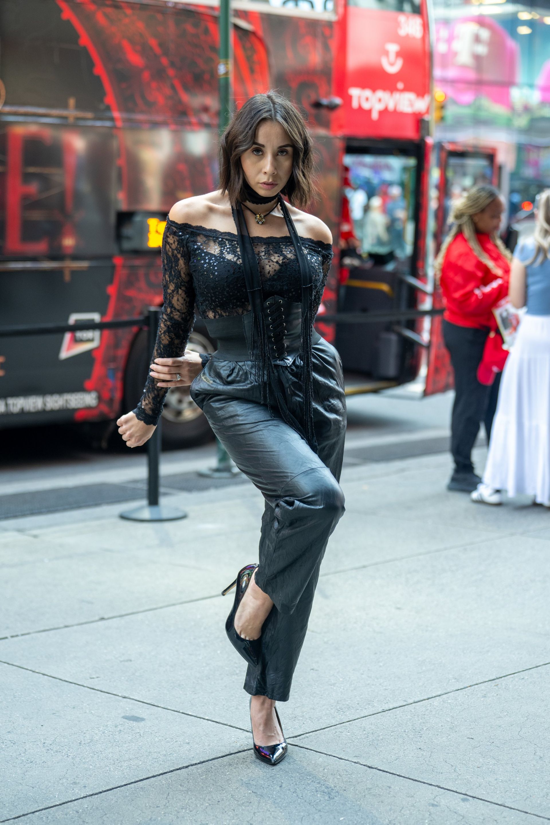 Woman posing on a city sidewalk in a black lace top and gray pants, with a red storefront in the background