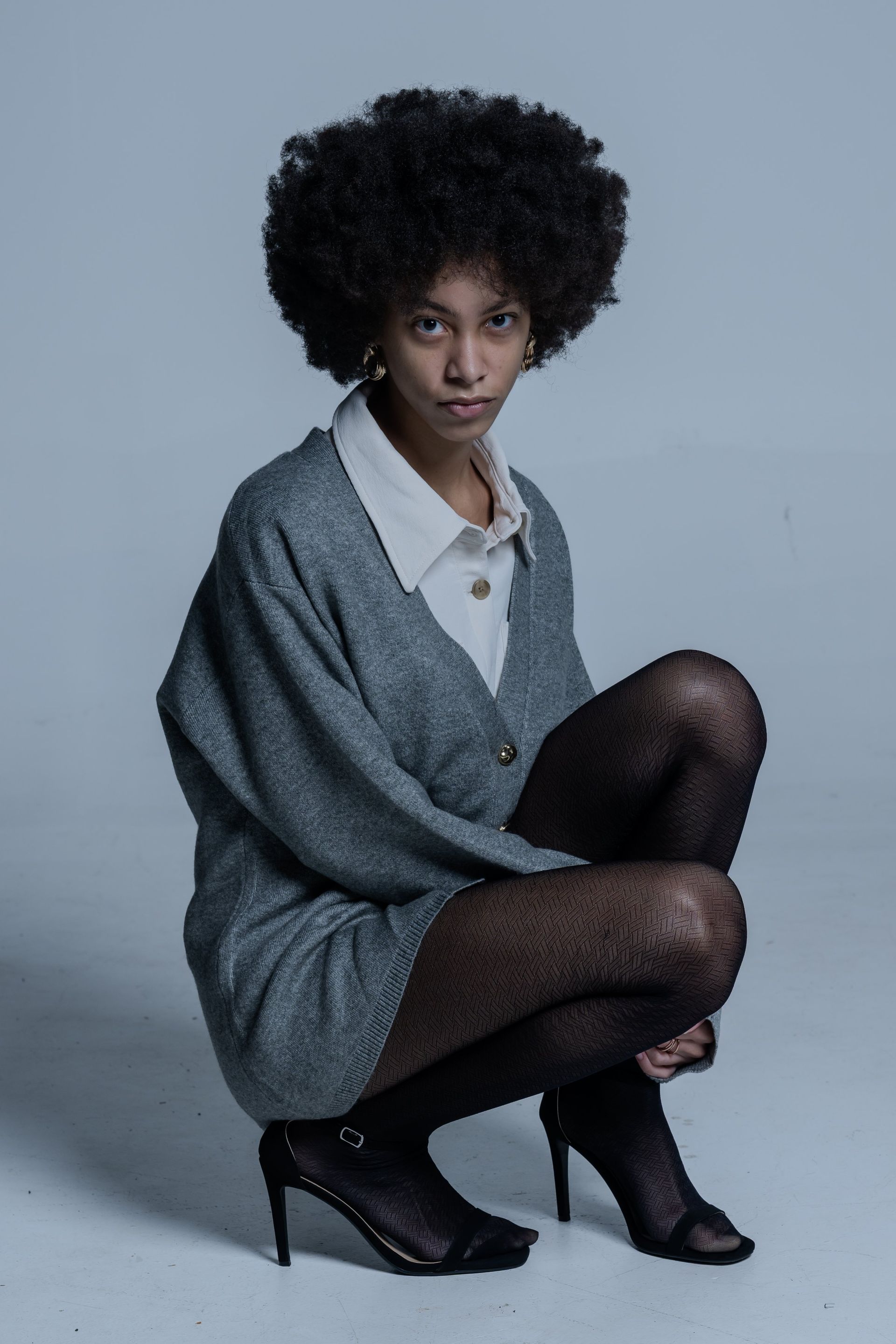 Person crouching in a gray oversized sweater, white collar shirt, black tights, and heels against a plain gray backdrop.