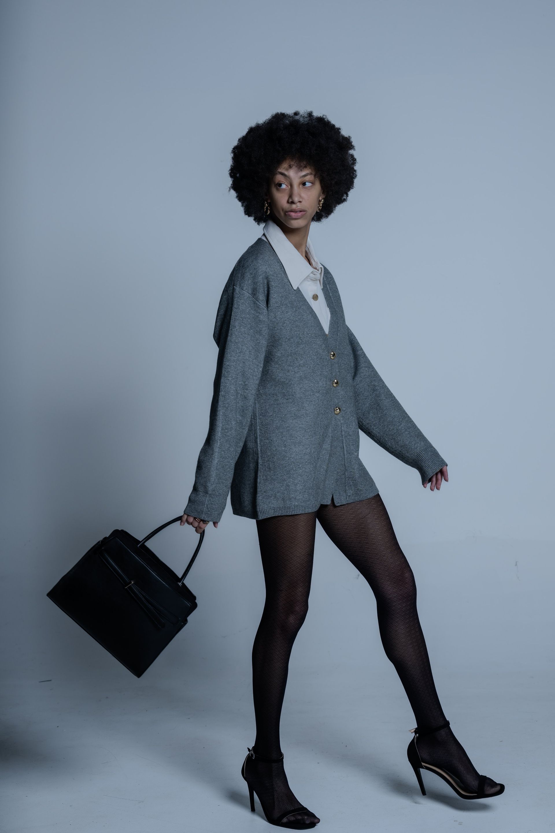 Woman in gray blazer and black tights walking with a black handbag against a plain background.