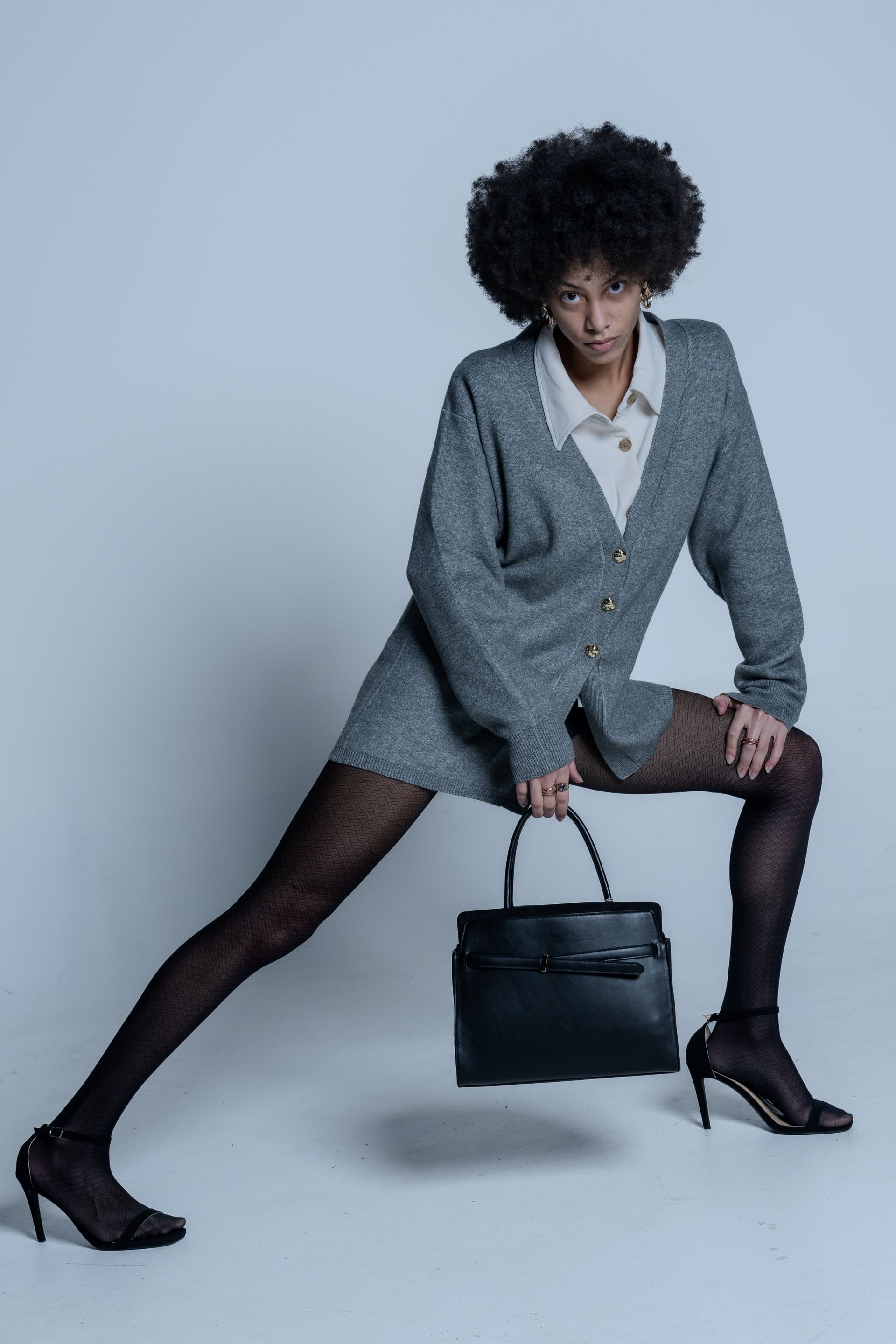 Fashion model posing in gray blazer and black heels, leaning with one leg extended and holding a black handbag.