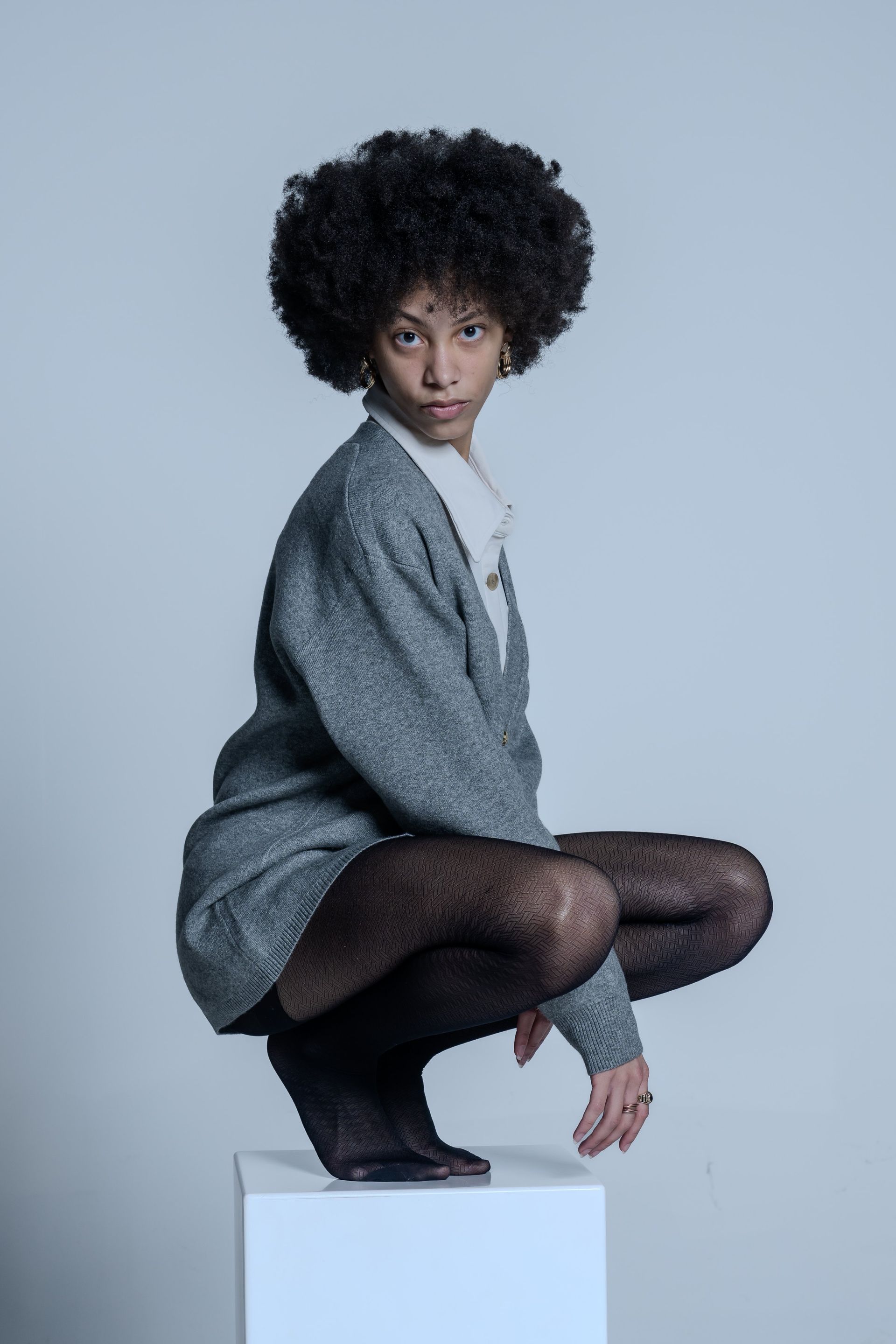 Woman posing in gray blazer and black tights on a white pedestal against a pale blue background
