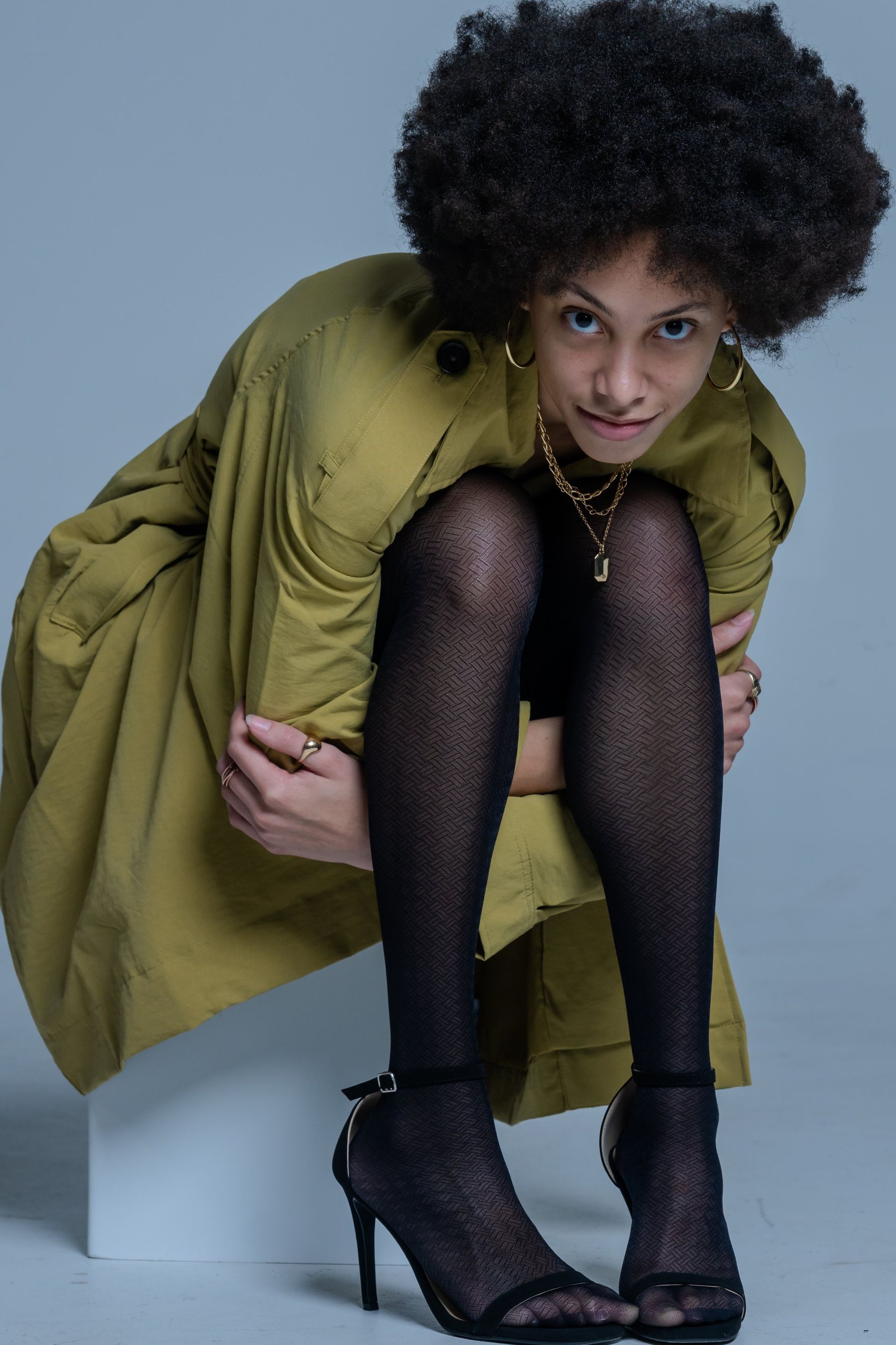 Person crouching in an olive coat and black tights, wearing black heels against a blue-gray background