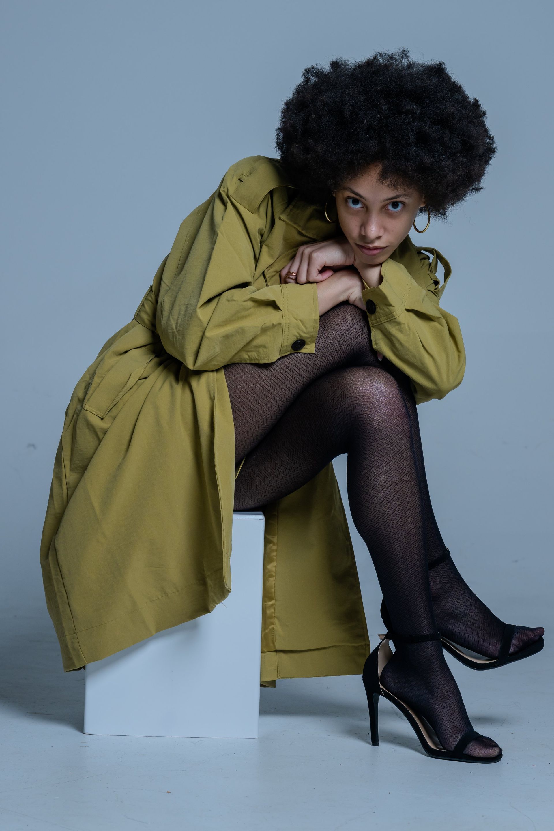 Model in olive coat and black tights poses seated on white blocks against a gray backdrop.
