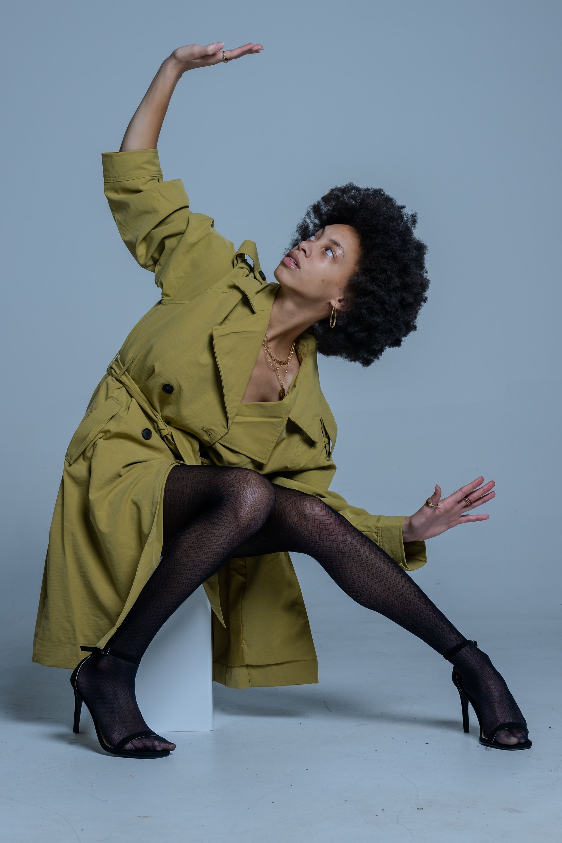 Model posing in an olive-green coat and black tights, seated low against a plain blue backdrop.